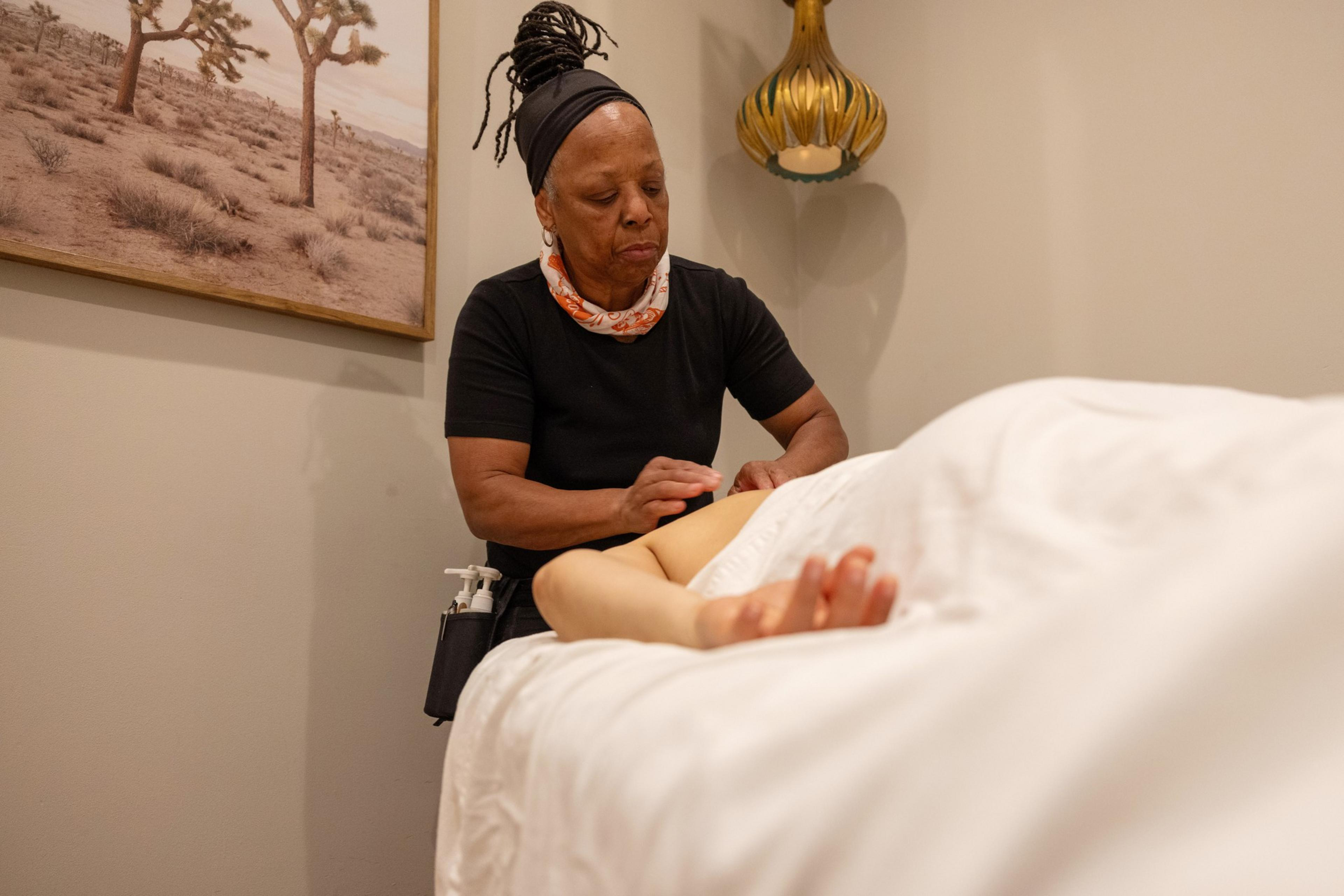 A person is giving a massage to someone lying on a bed, covered with a white sheet. The room has a desert landscape painting and a decorative lamp on the wall.