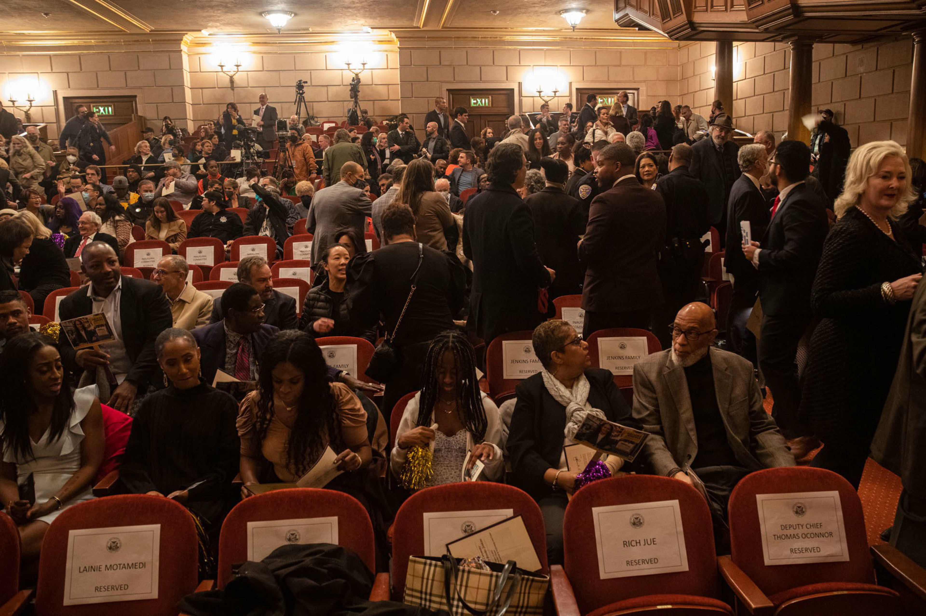 A crowded auditorium with people sitting and standing, reserved seats with name tags, and a lively atmosphere before an event.