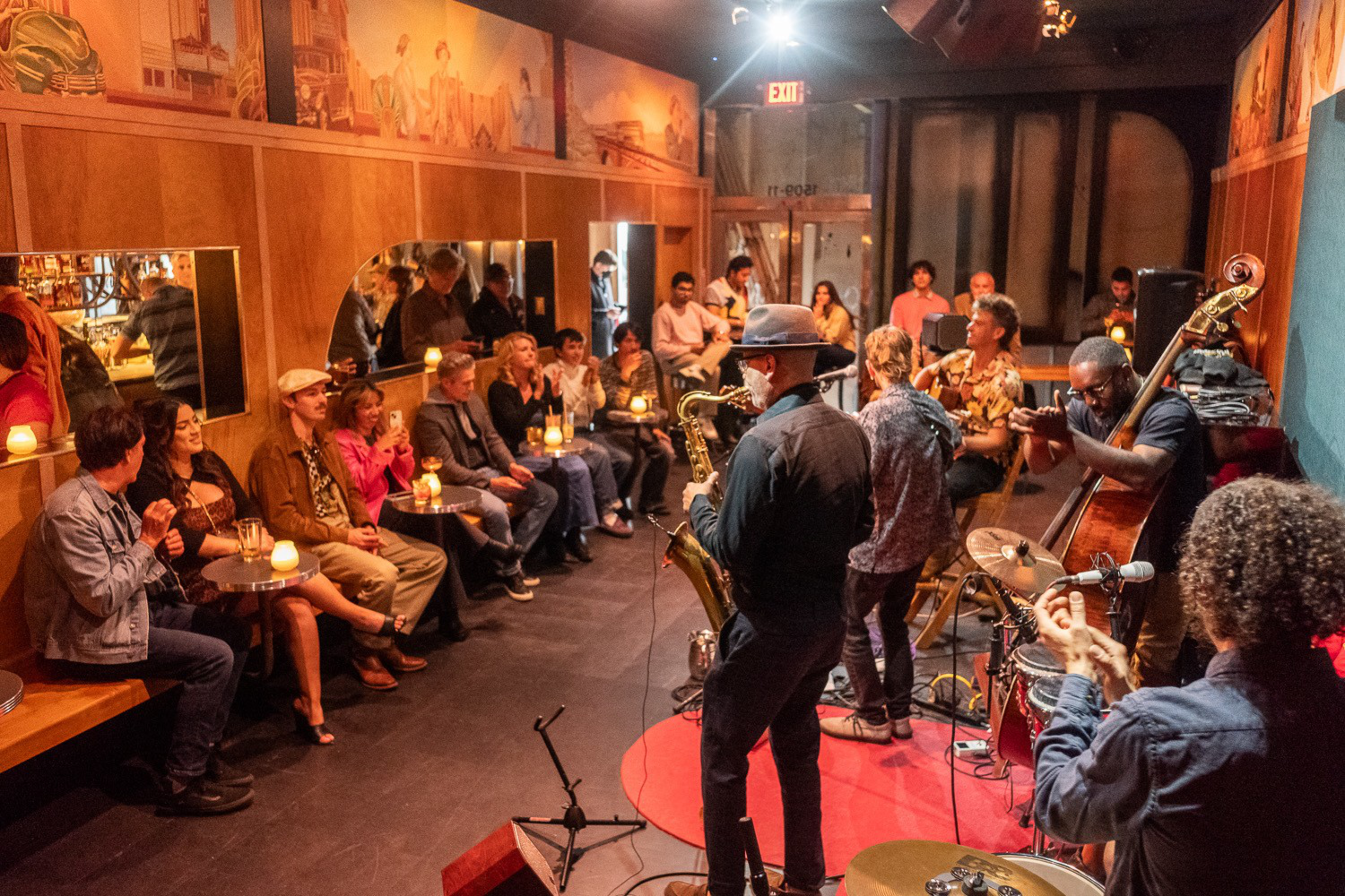 A jazz band performs on a small stage while a diverse audience sits closely together, watching and enjoying the music in a cozy room.