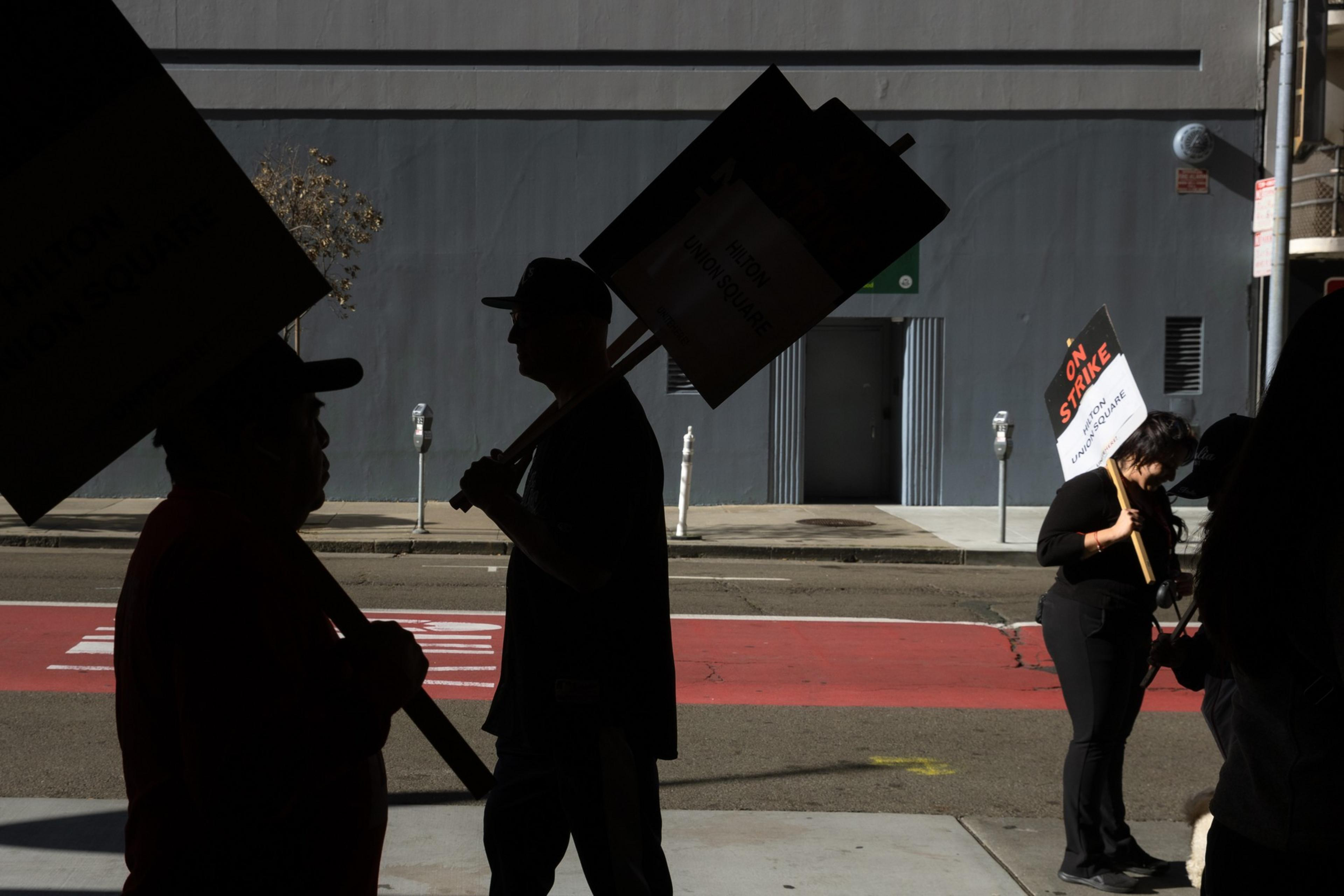 People carrying &quot;On Strike&quot; signs walk along a sidewalk, their silhouettes stark against the backdrop of a gray building and an empty street.
