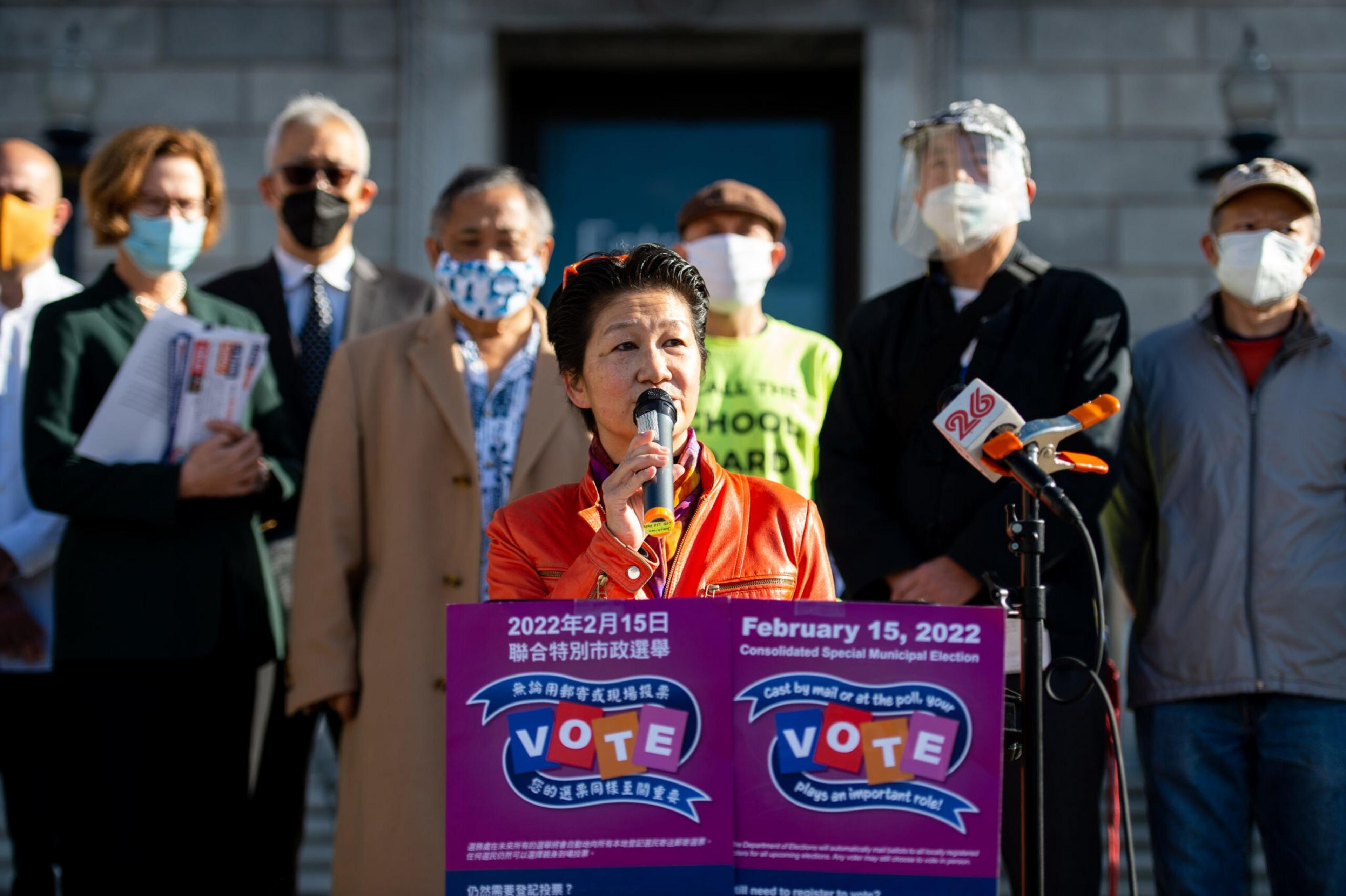 A woman in a red jacket speaks at a podium with bilingual election signs, surrounded by masked and shielded people, some holding papers.