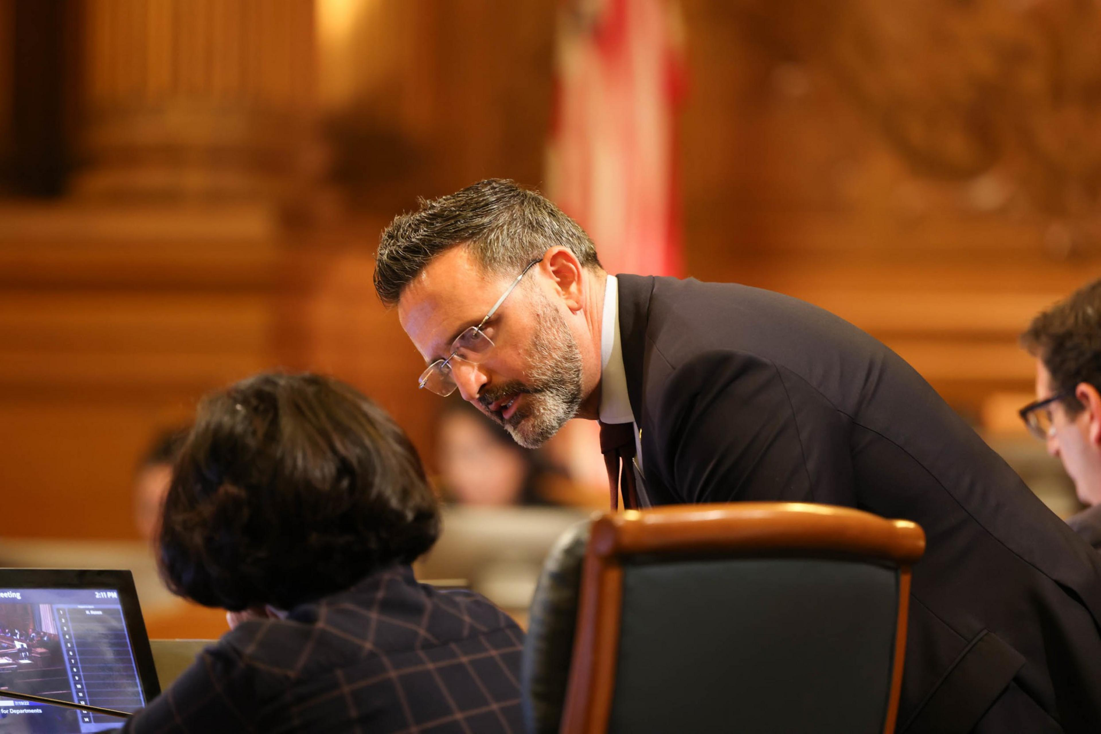 A man in a suit leans over to speak with a seated woman working on a laptop in a formal setting with an American flag blurred in the background.