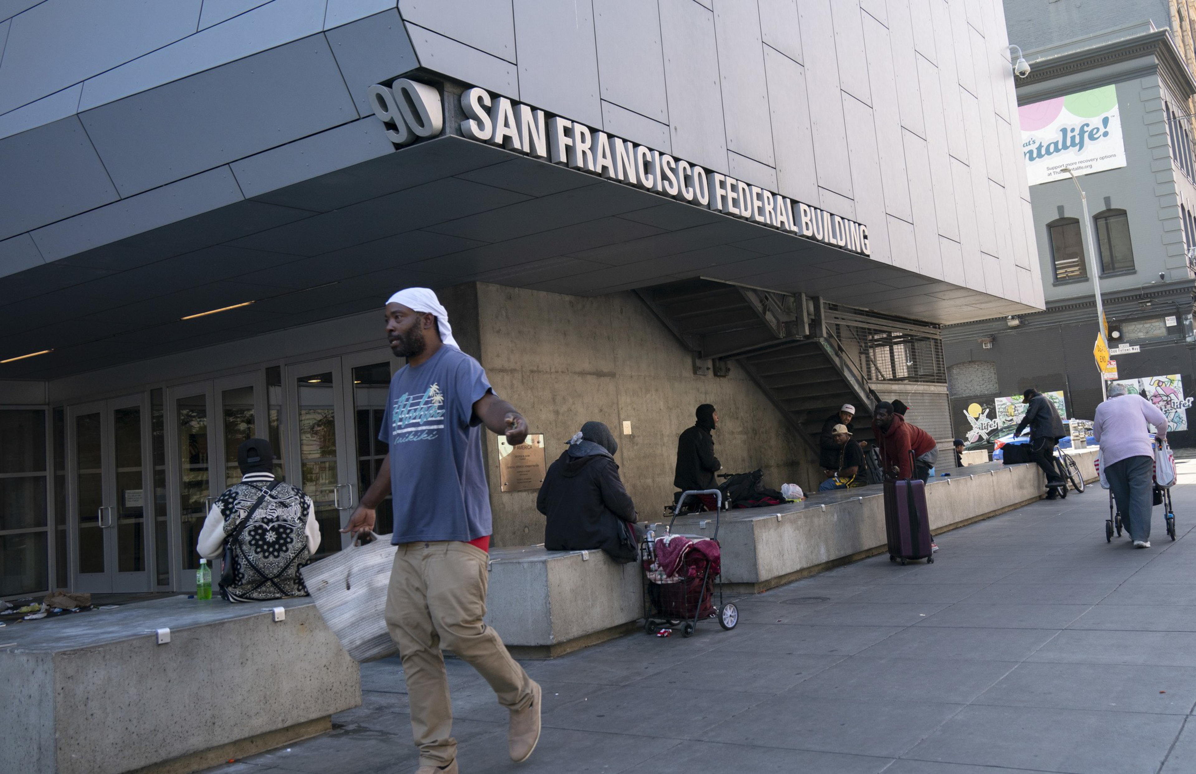 The image shows a group of people outside the San Francisco Federal Building. Some are sitting or standing with belongings, such as carts and bags, on a concrete bench.