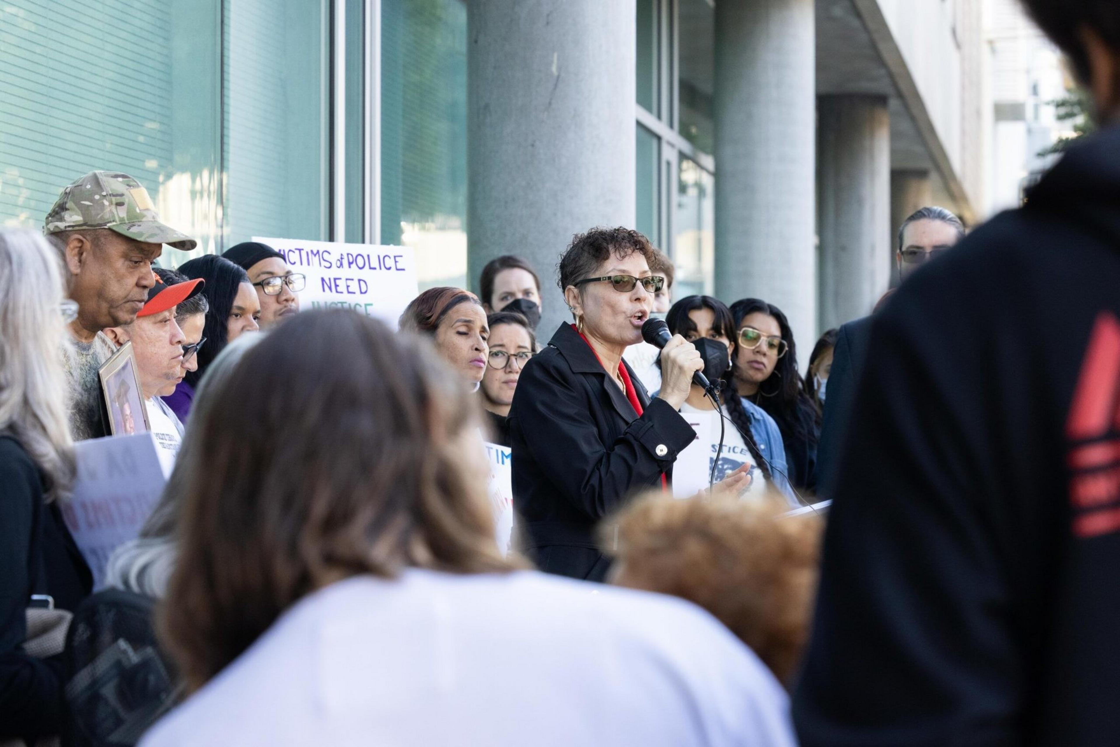 A woman speaks into a microphone addressing a crowd holding signs that say "Victims of Police Need Justice" outside a building with glass windows.