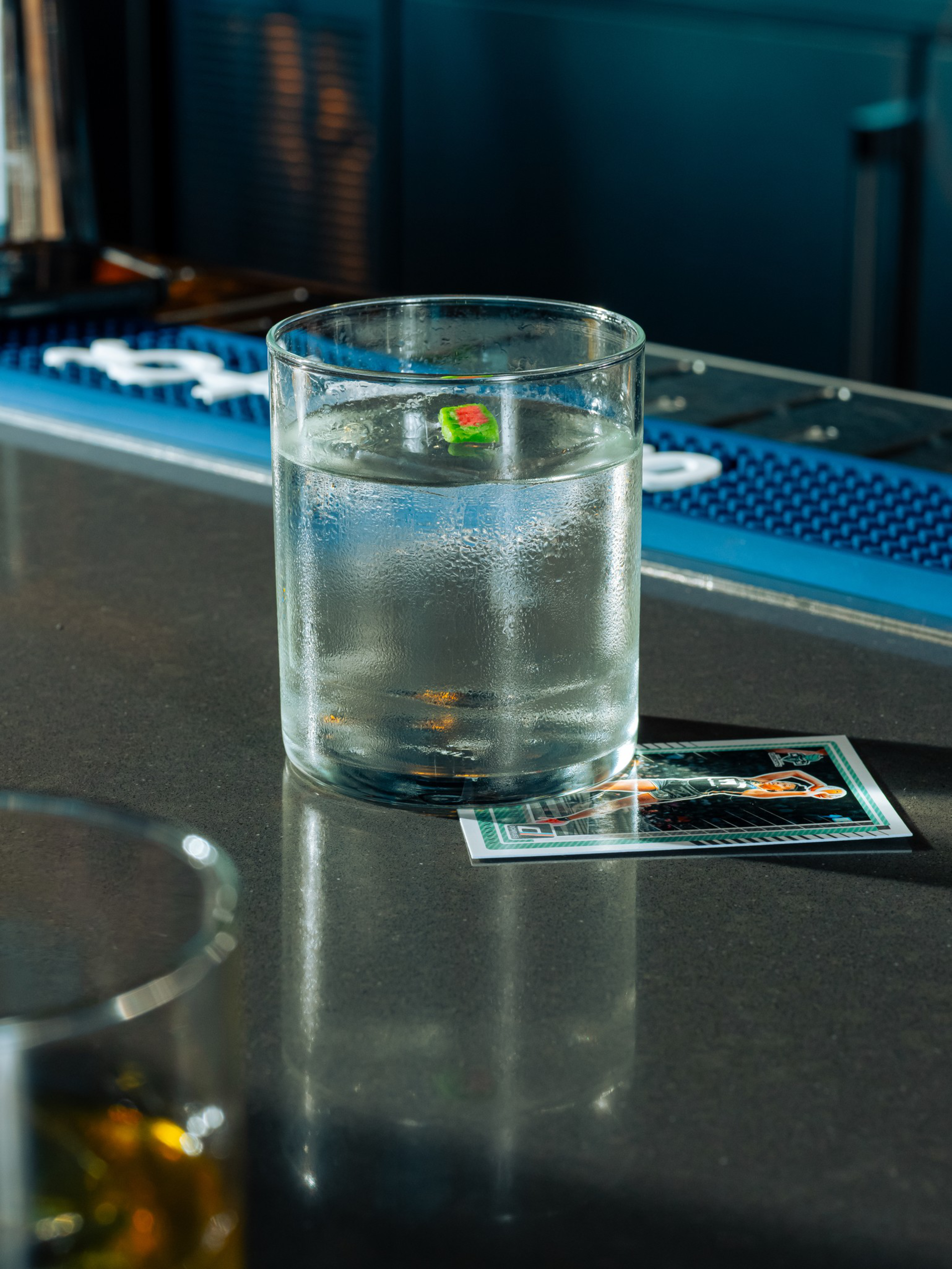 A clear glass of water with condensation sits on a bar counter, partly covering a playing card, with another drink blurred in the foreground.