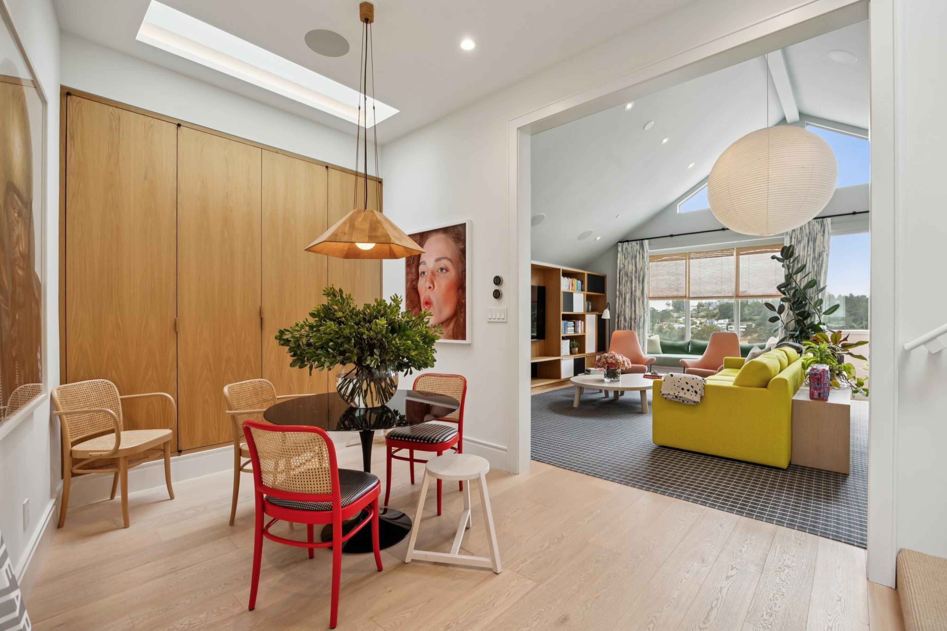 A dining area with a round table, mixed chairs including two red ones, a plant centerpiece, wooden floors, and an adjacent living room with a yellow sofa and large windows.