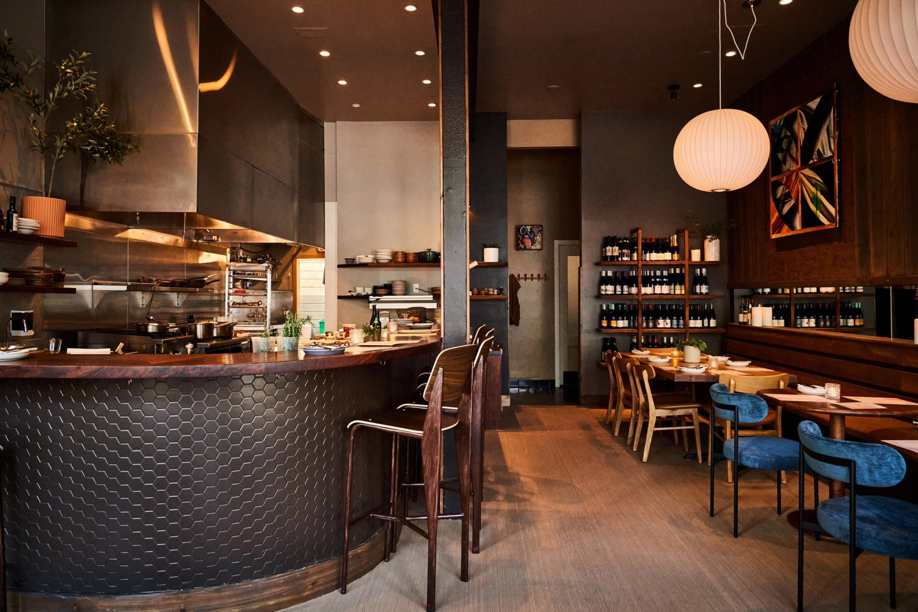 The image shows a cozy restaurant interior with a hexagonal-tiled bar, wooden stools, round pendant lights, shelves of wine, and neatly set tables.