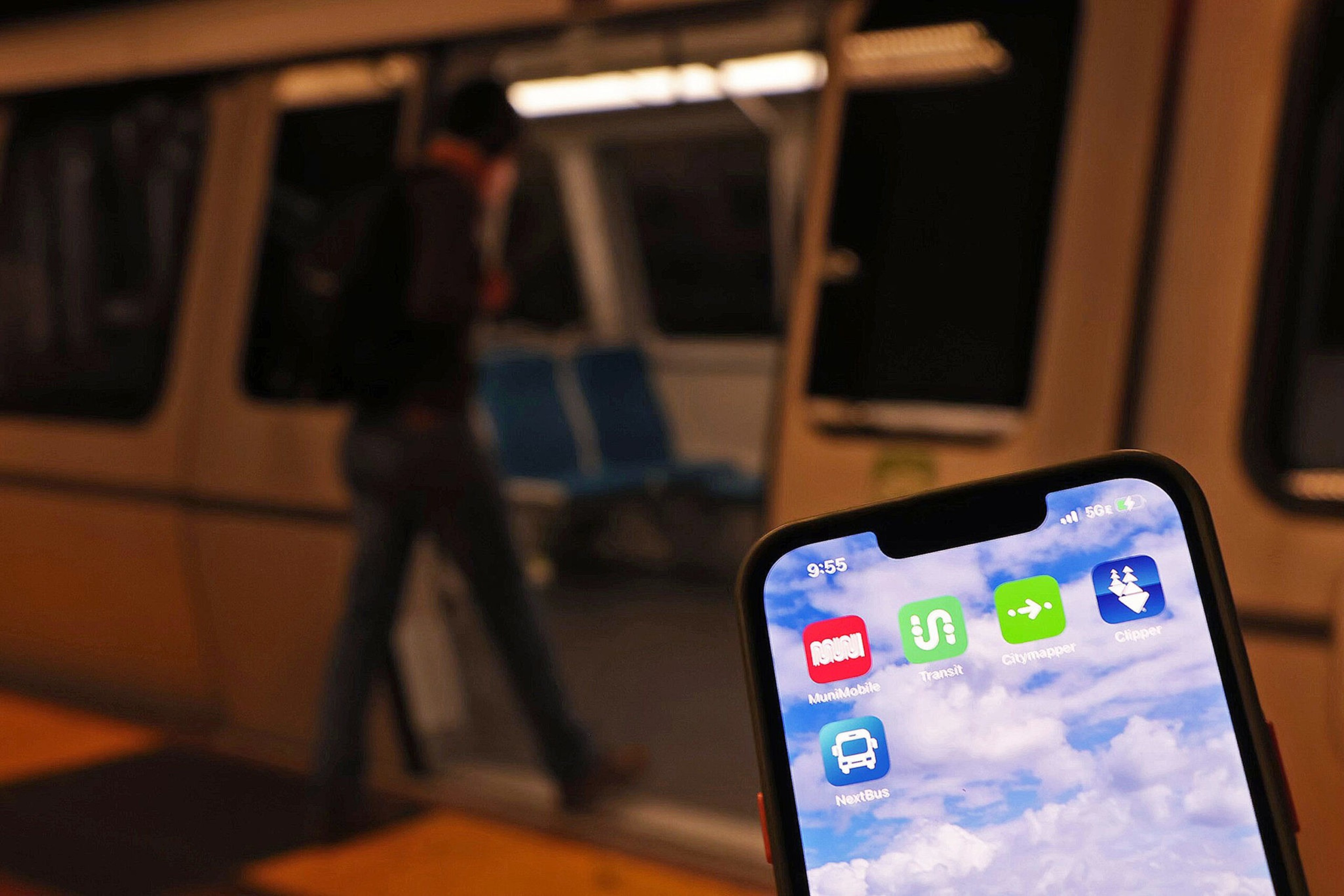 A hand holds a smartphone displaying transit apps, with a blurred person boarding a subway train in the background.