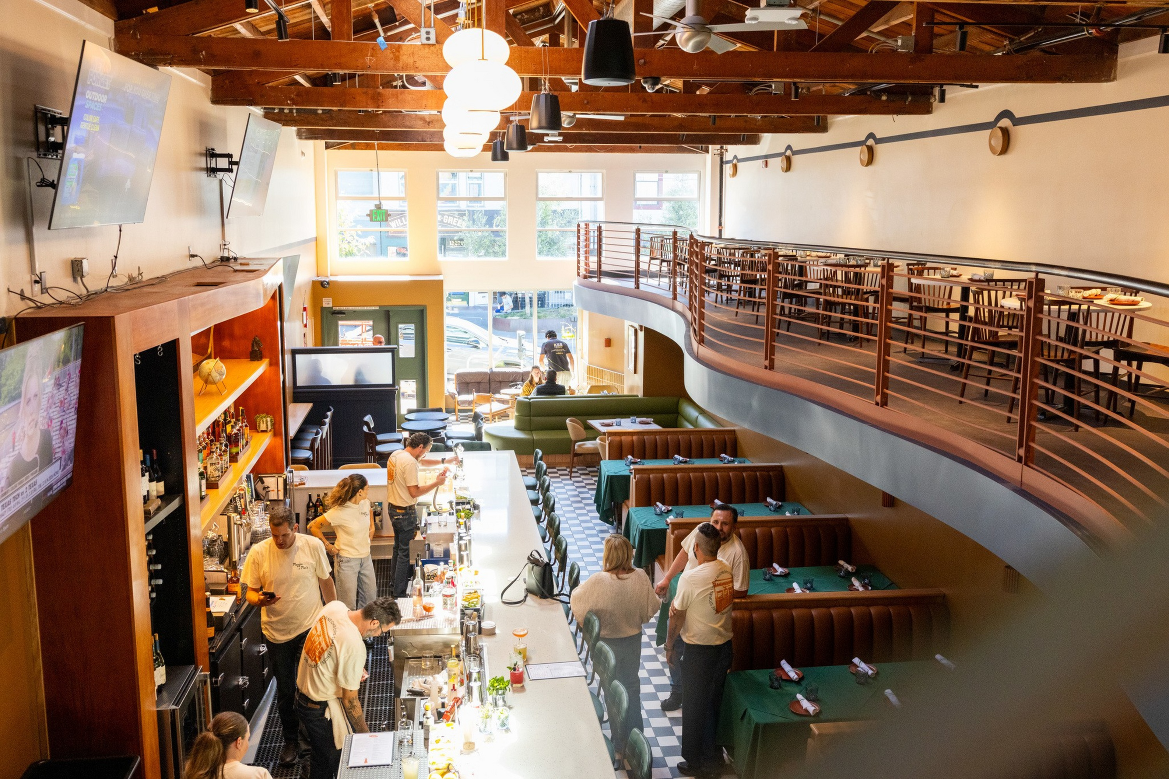 A bright restaurant interior with wooden beams, a long bar with patrons and staff, green booth seating, and an upper-level dining area with tables and chairs.