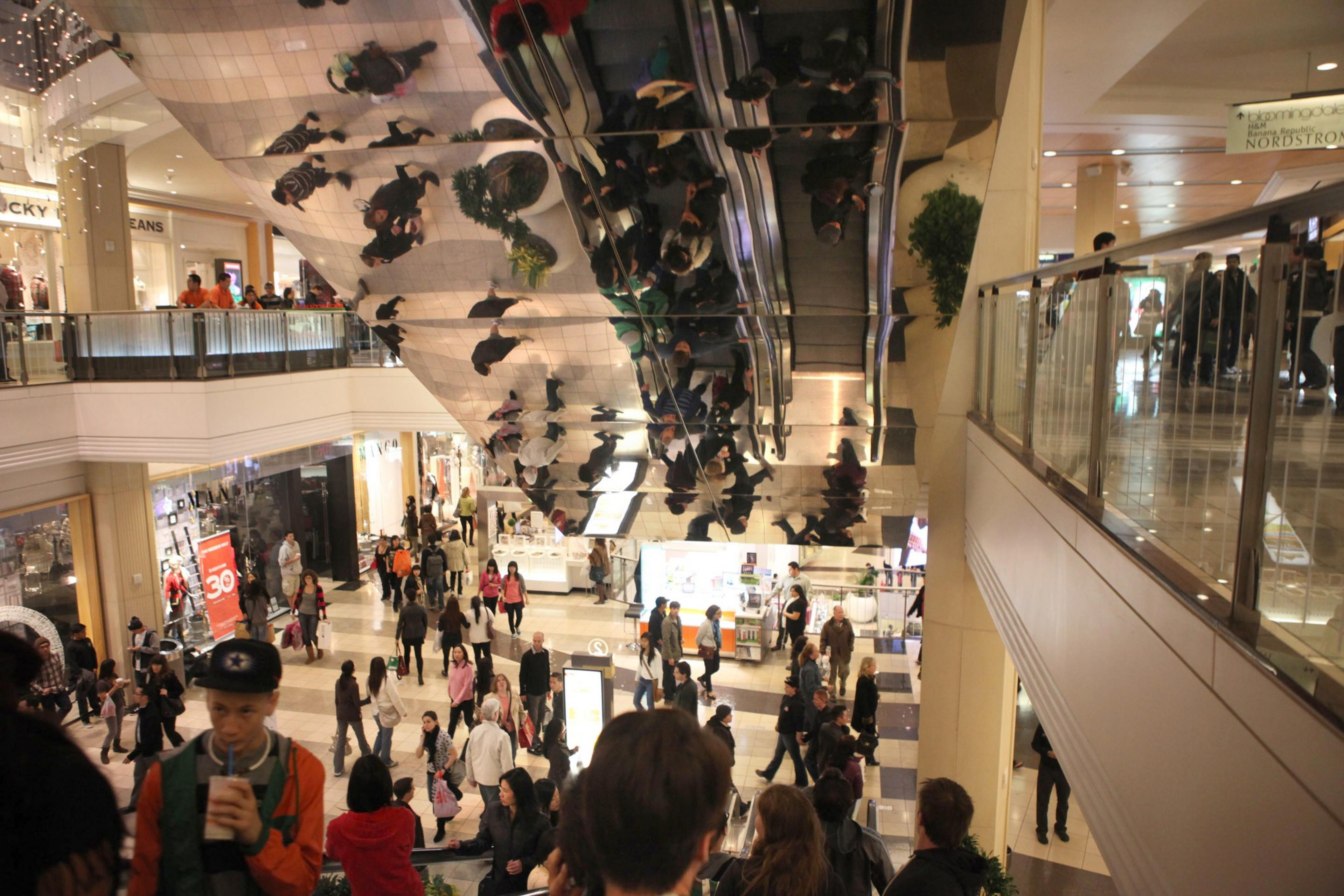 A bustling mall interior with shoppers and reflective ceiling doubling images of people and storefronts.