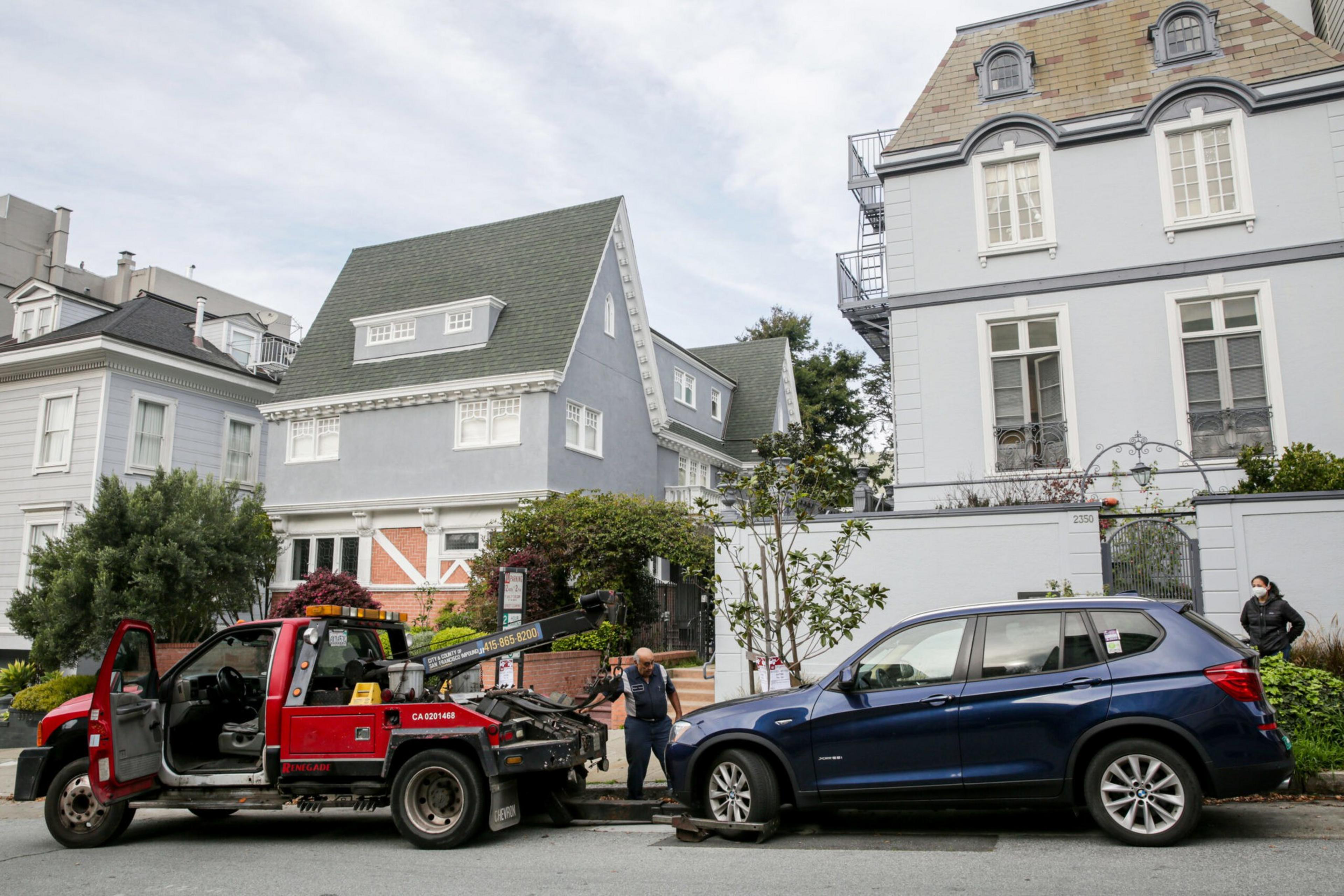 A red tow truck is towing a blue SUV parked on a street in front of gray houses. A person is operating the tow, and another person stands nearby.