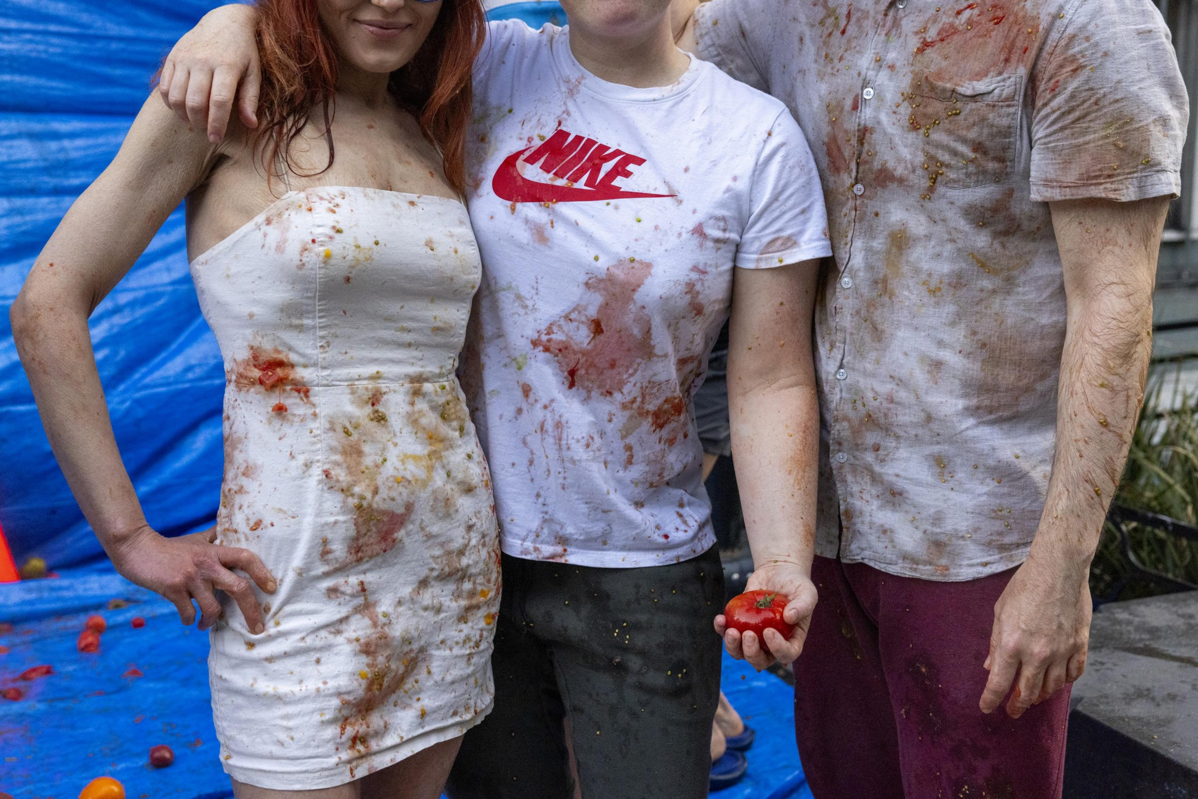 Three people, each wearing thoroughly soiled clothing, pose in front of a blue tarp, likely after a messy event involving tomatoes, as one holds a tomato in hand.