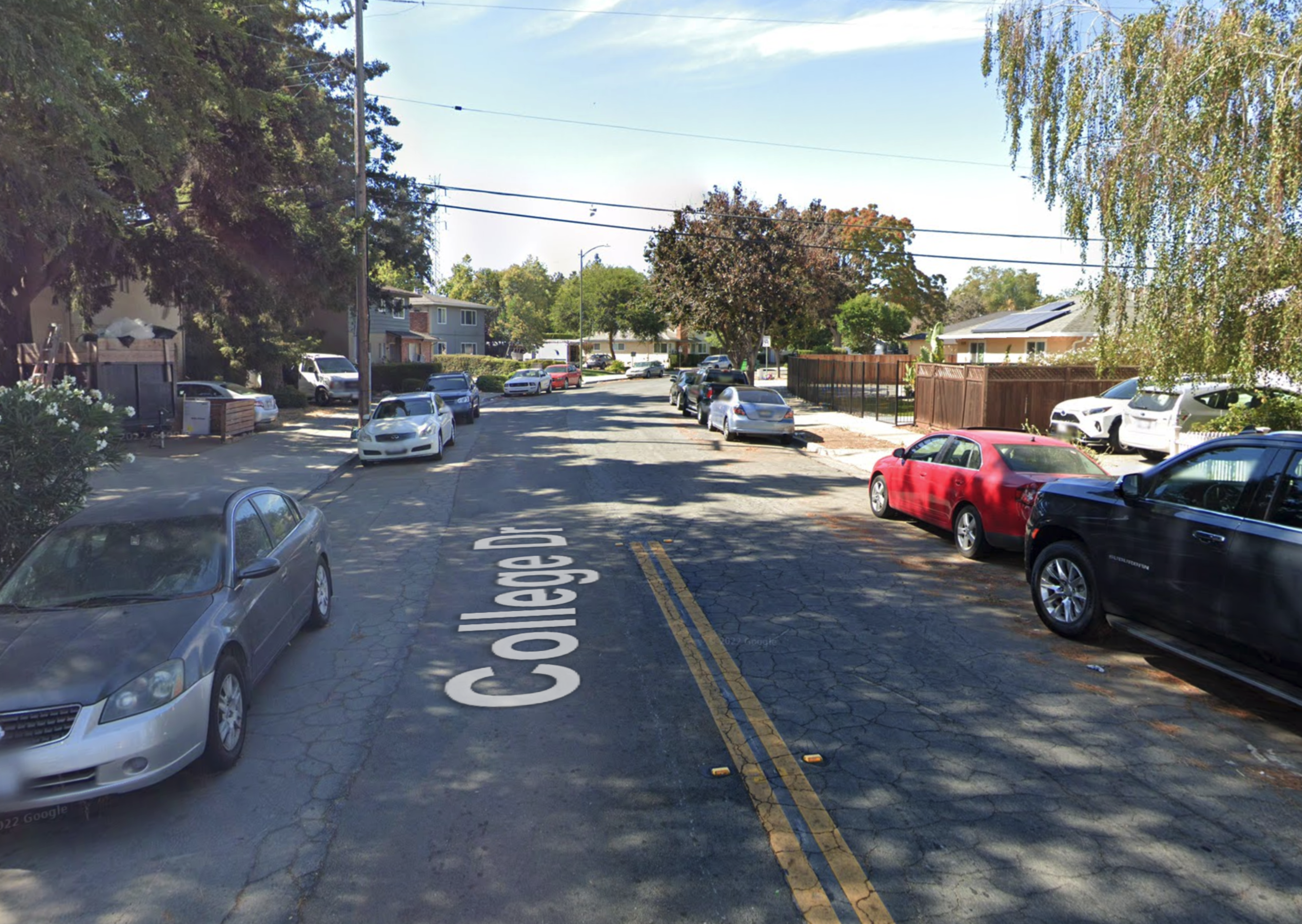 A quiet residential street named College Dr has cars parked on both sides and houses with trees and fences lining the road under a partly cloudy sky.