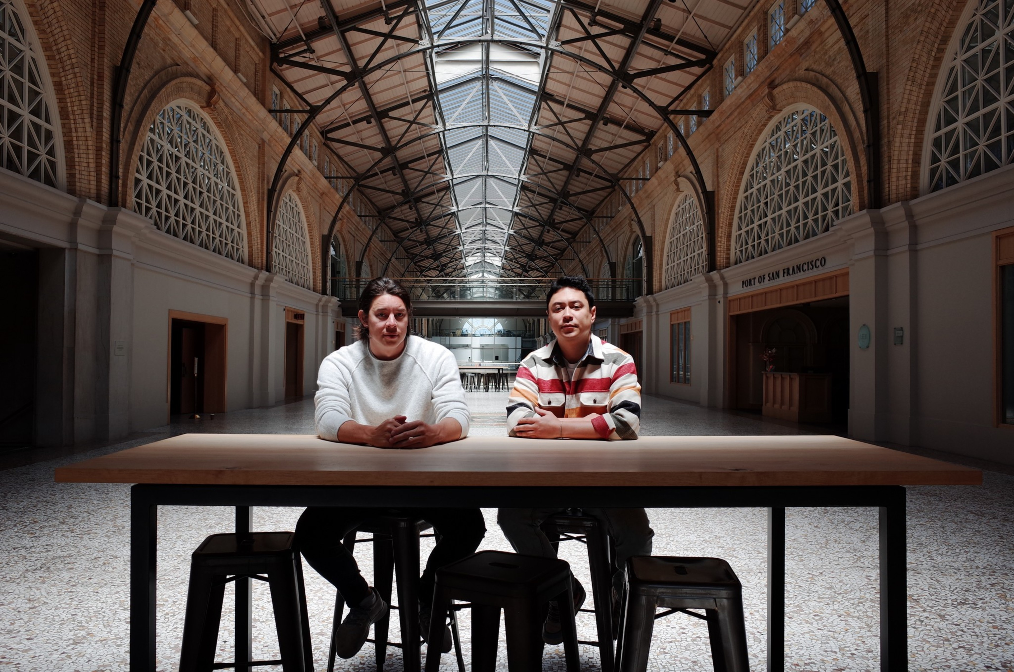 Two men sit at a wooden table with stools in a large, sunlit hall featuring arched brick walls, a glass ceiling, and a sign reading “Port of San Francisco.”