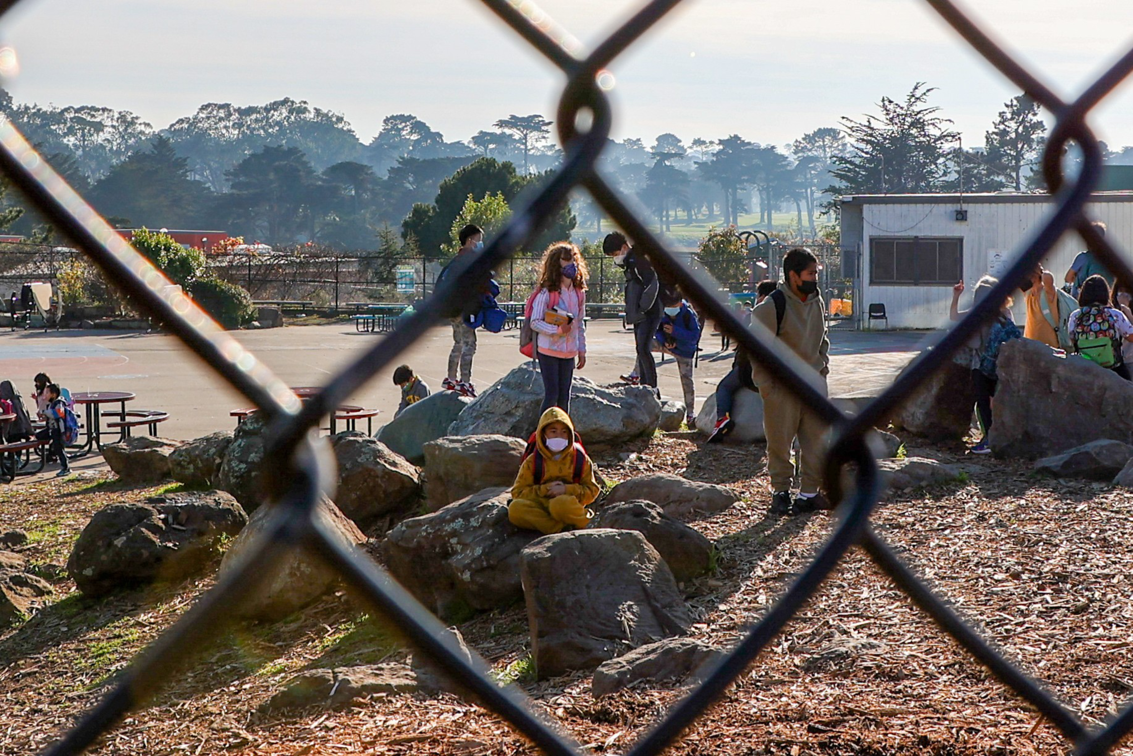 Children wearing masks play and sit on large rocks in a schoolyard, surrounded by a chain-link fence on a sunny day.