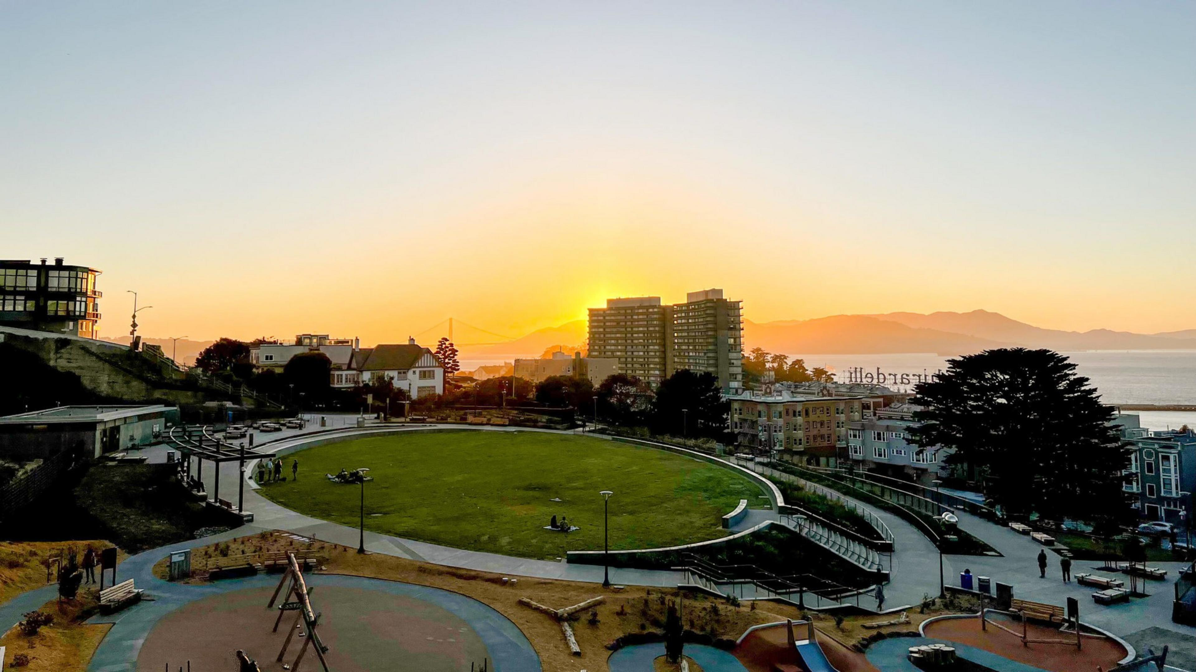 A sunset cityscape with Francisco Park seen at sunset with buildings and distant mountains in the background under a golden sky.