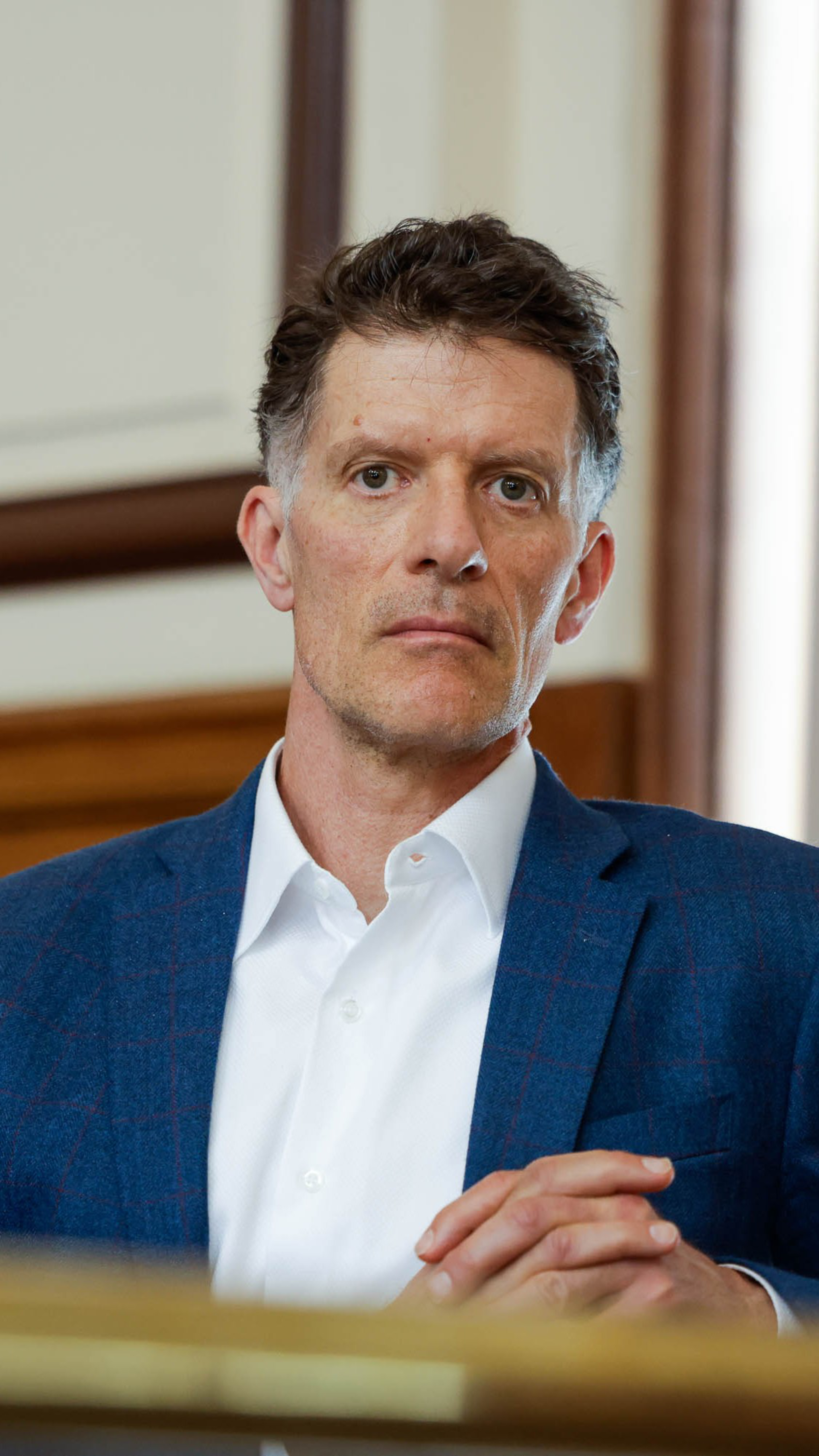 A man in a blue blazer and white shirt, with a serious expression, sits with hands clasped in front of him.