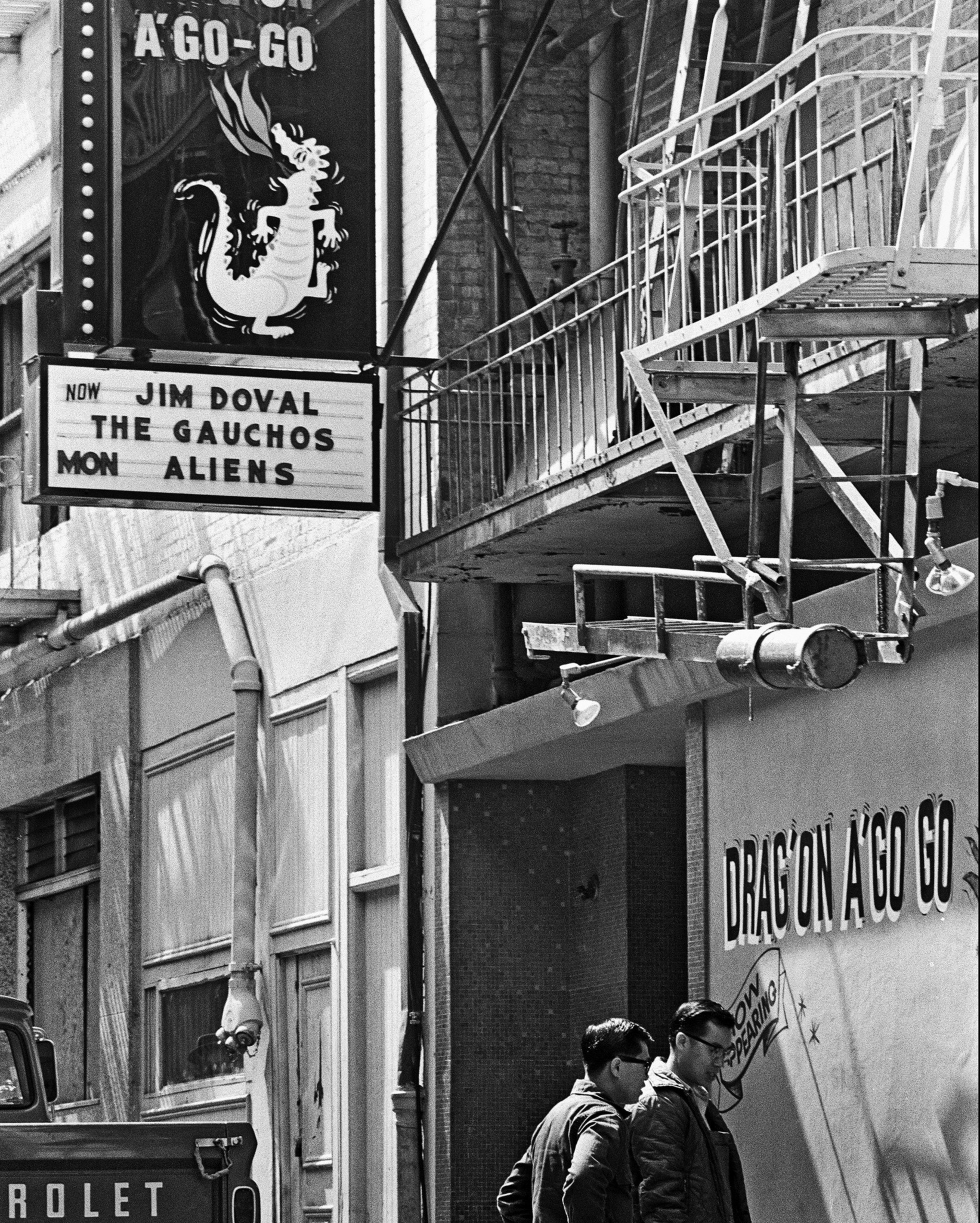 A black-and-white photo of a street corner with a "Dragon A Go-Go" sign, fire escape stairs, and two men walking by, one looking at the sign.