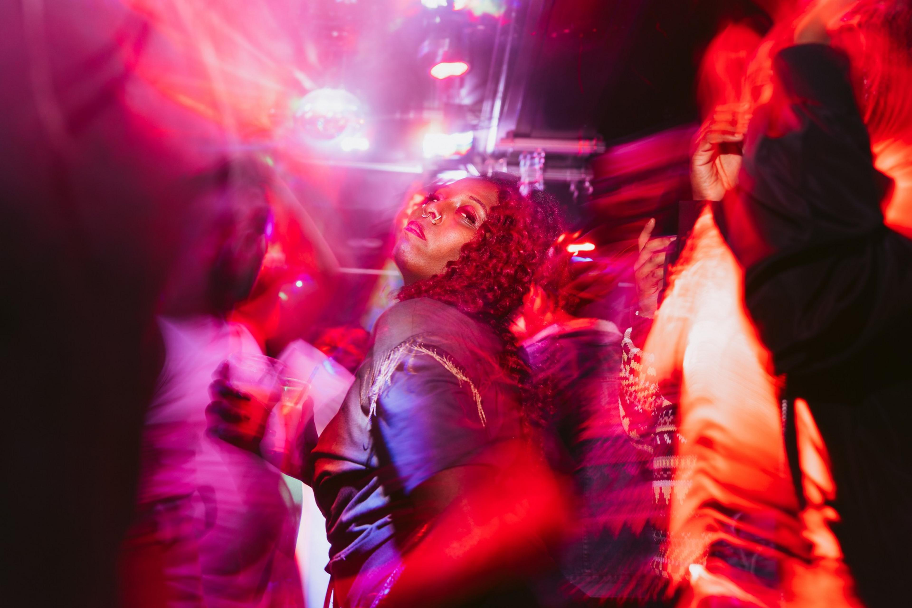 A person with curly hair and a nose ring is in a vibrant, colorful nightclub scene, surrounded by blurred figures and glowing red and pink lights.