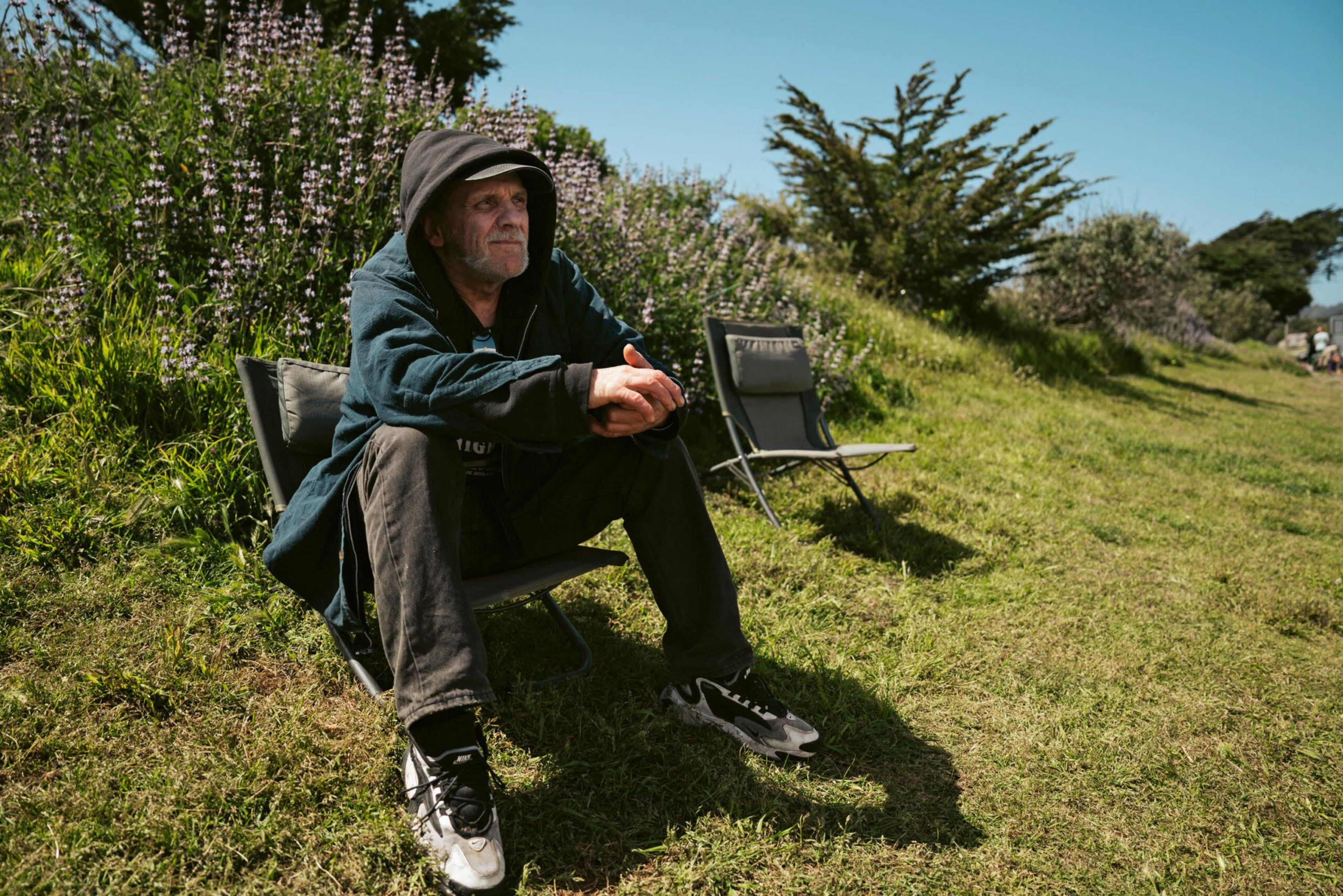 An older man in a hooded jacket sits on a low chair on grass, with purple flowers and plain chairs behind him under a sunny sky.
