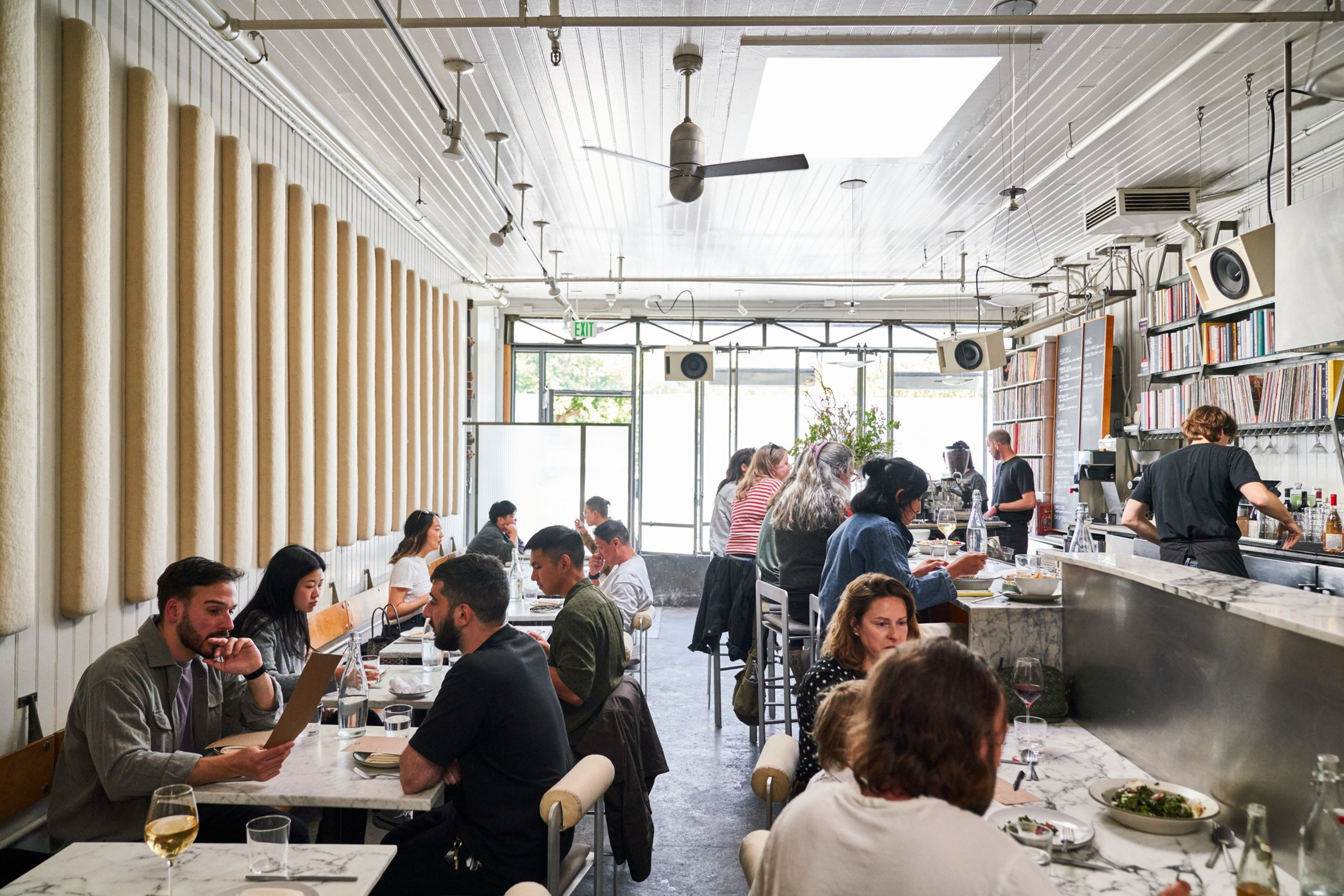 The image shows a bustling, modern cafe with people dining and conversing. There are long marble tables, a bar area, and a wall lined with records.