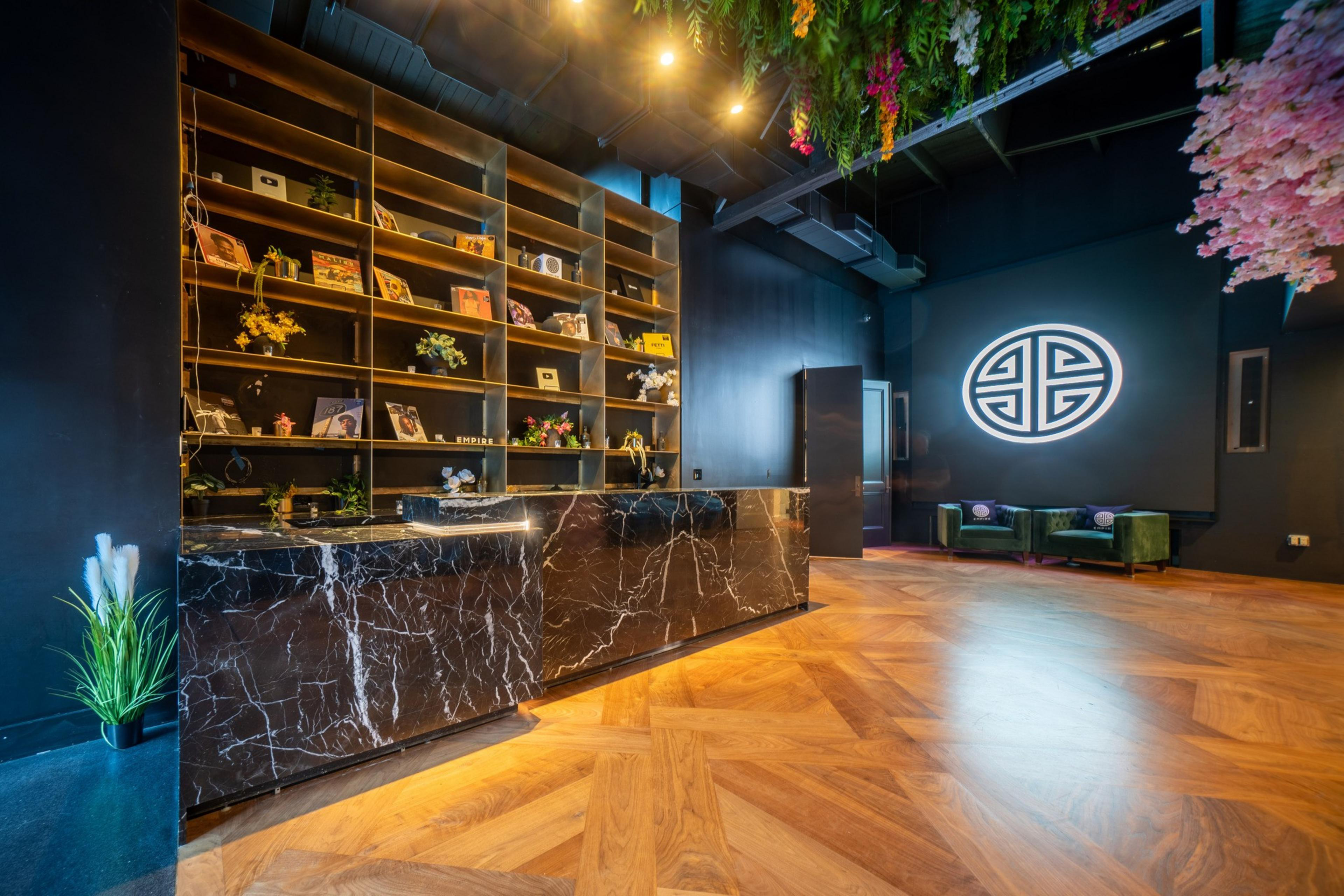 The image shows a modern room with a black marble reception desk, wooden flooring, and a high ceiling with hanging plants. Shelves display books and decor.