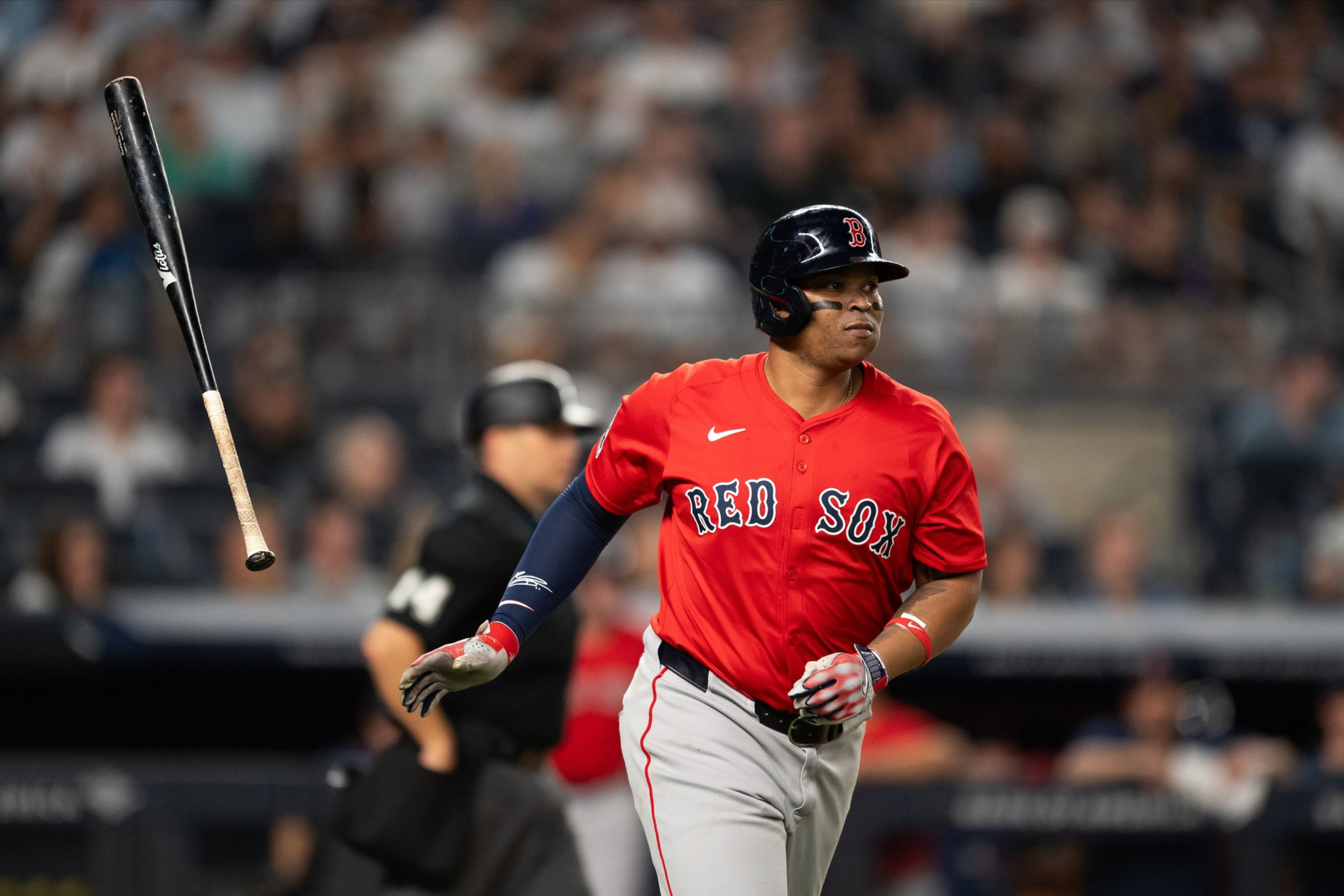 A baseball player in a red &quot;Red Sox&quot; jersey watches his hit, helmet on, with a bat mid-air, and a blurred crowd behind him.
