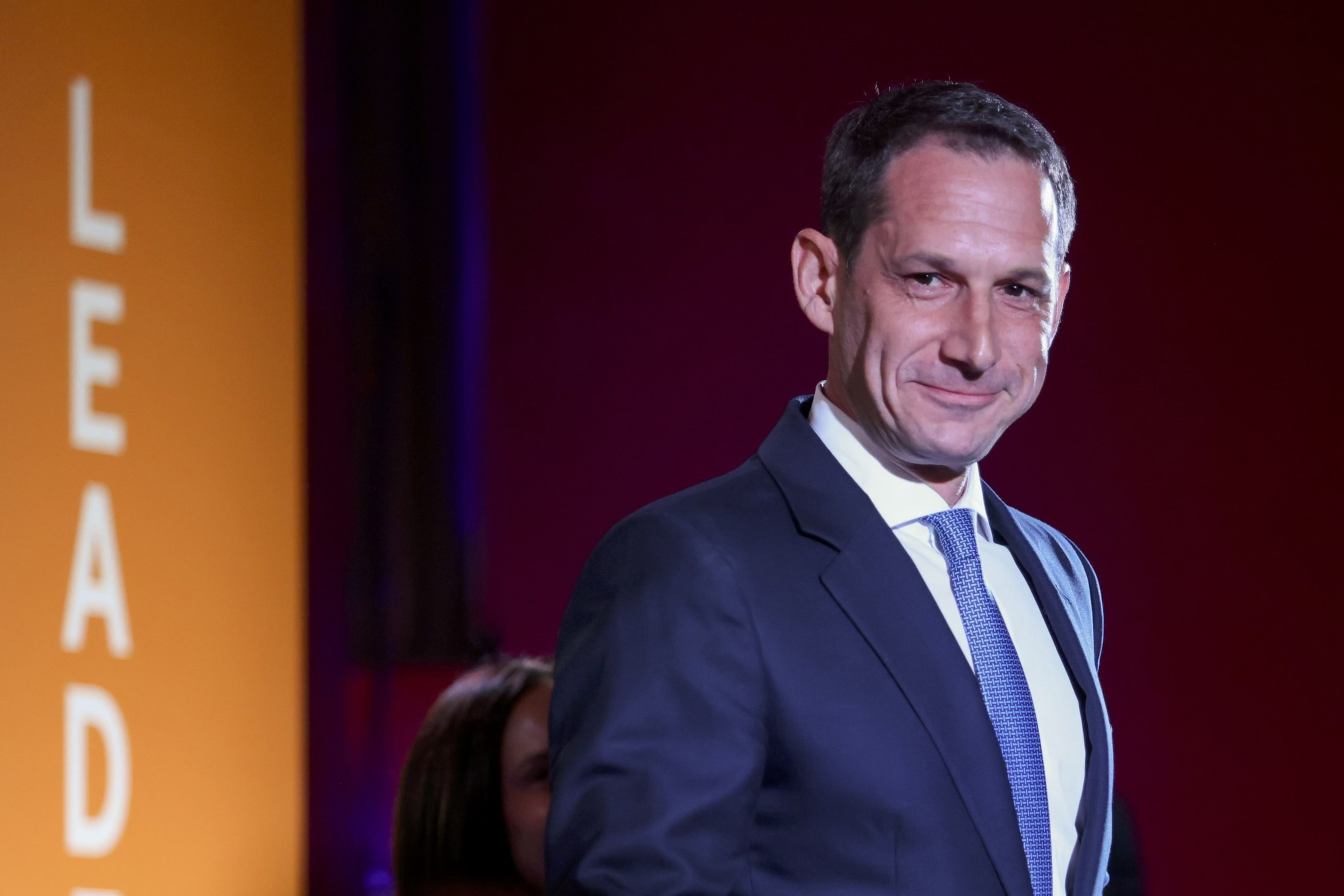 A man in a suit and blue tie smiles slightly while standing in front of a glowing orange backdrop with the word "LEADER" partially visible.
