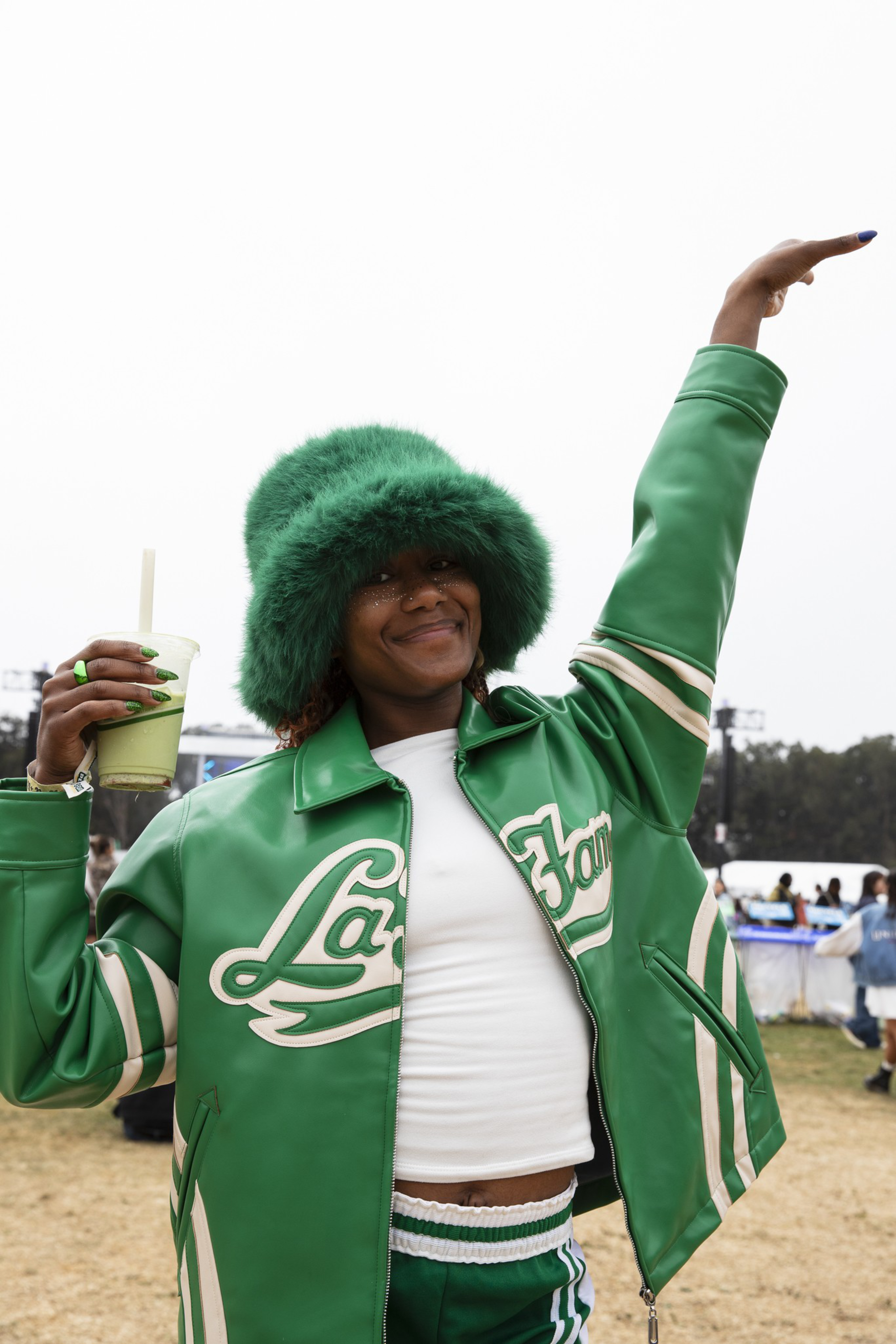 A person wears a green furry hat, green jacket with white lettering, and matching pants, holding a green drink and smiling with one arm raised outdoors.