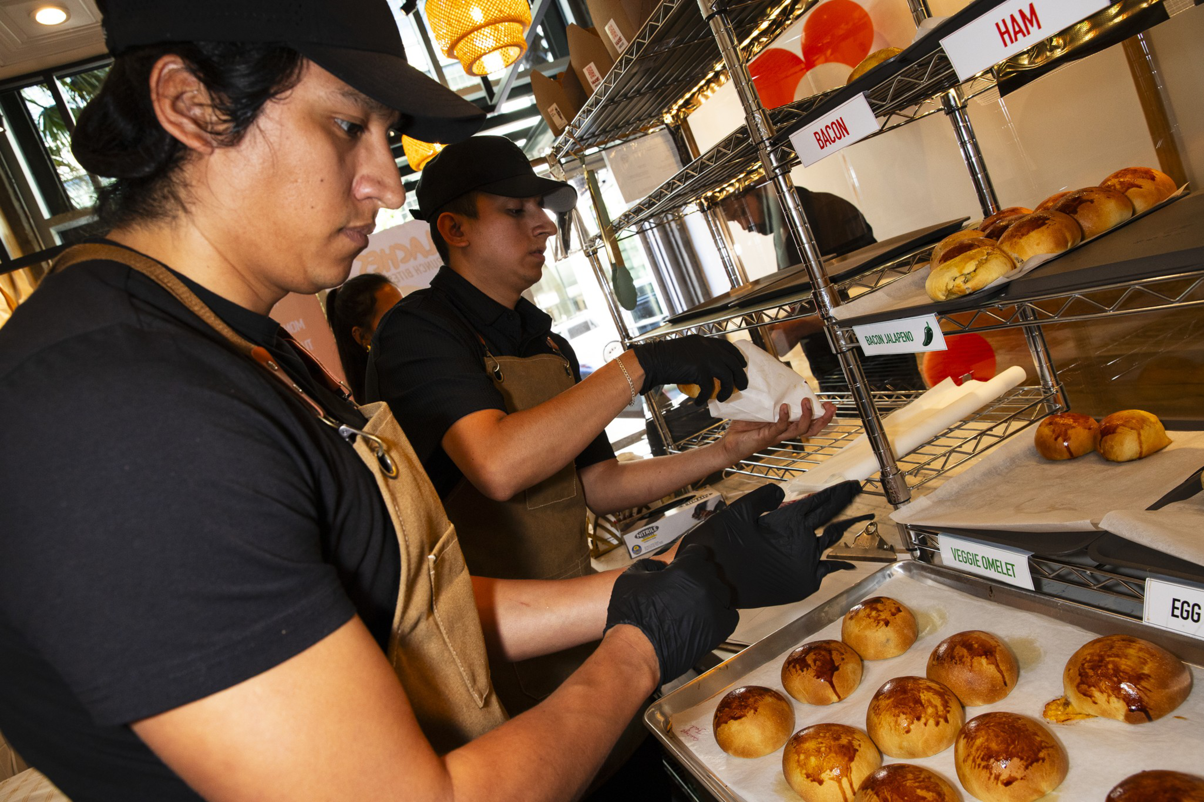 Two workers wearing black shirts, black gloves, and brown aprons packaging freshly baked rolls labeled by different fillings like bacon, ham, and veggie omelet.