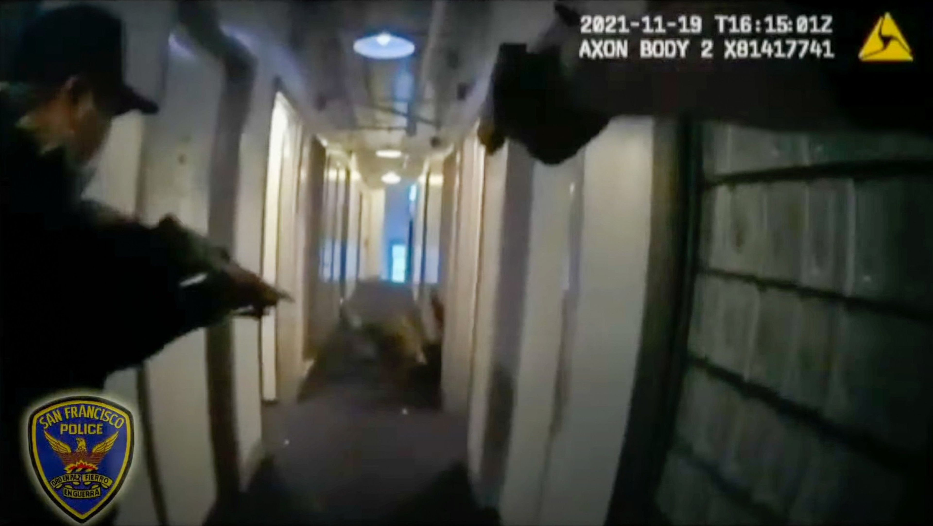 A police officer, seen from behind, aims a weapon down a narrow, well-lit corridor. The San Francisco Police badge is visible in the lower corner.