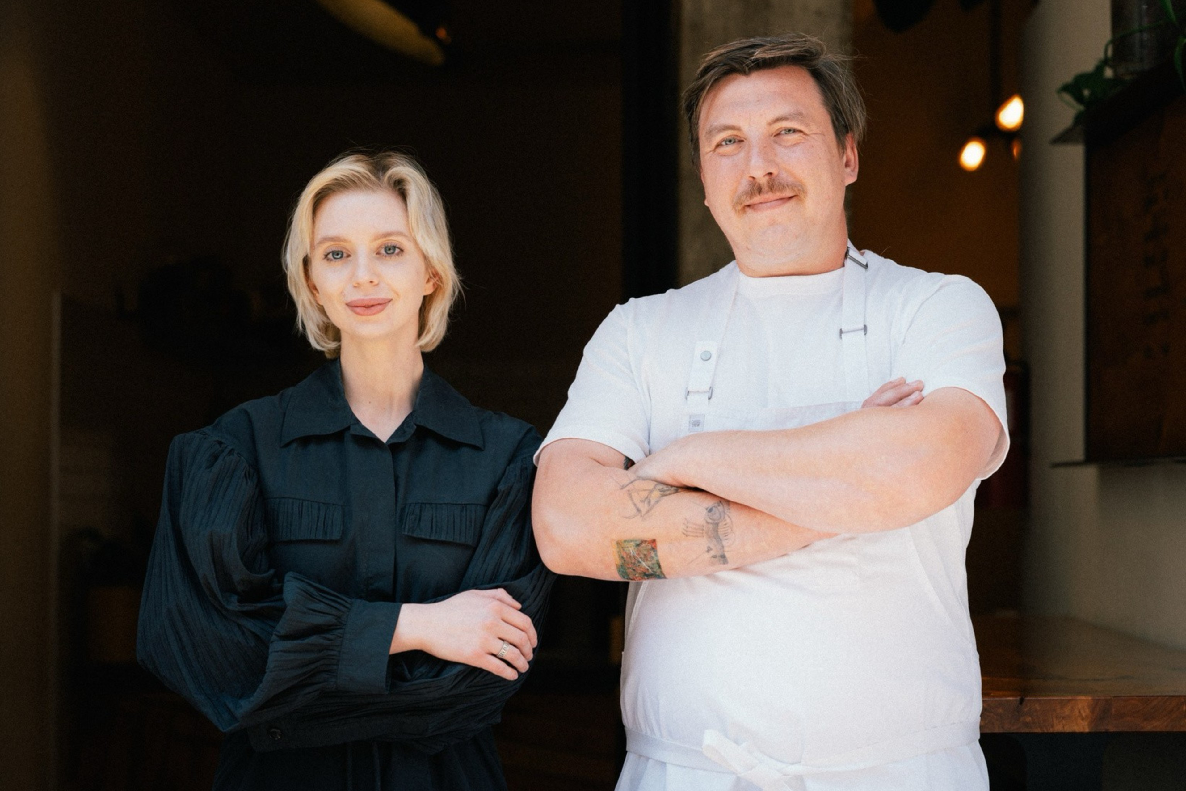 A man in a white shirt and apron stands beside a woman in a black dress. They're in a dimly lit space with light fixtures above them, appearing calm and composed.