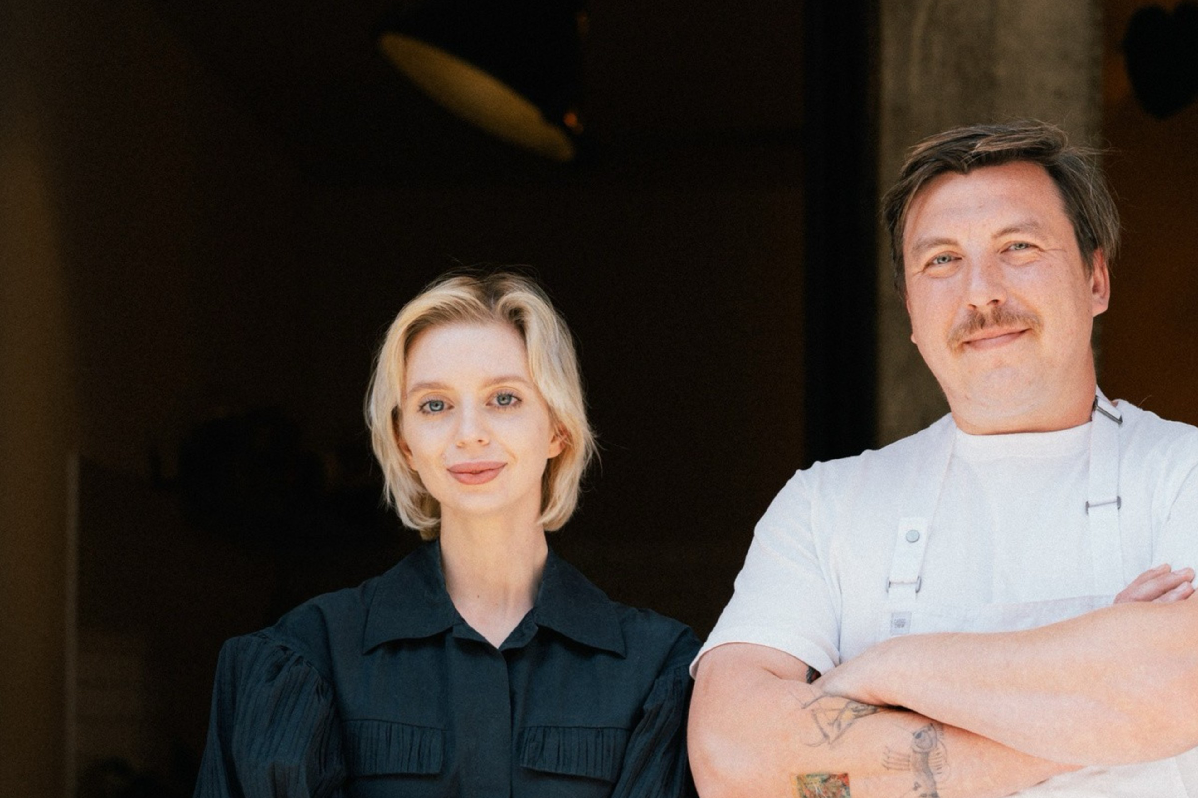 A man in a white shirt and apron stands beside a woman in a black dress. They're in a dimly lit space with light fixtures above them, appearing calm and composed.