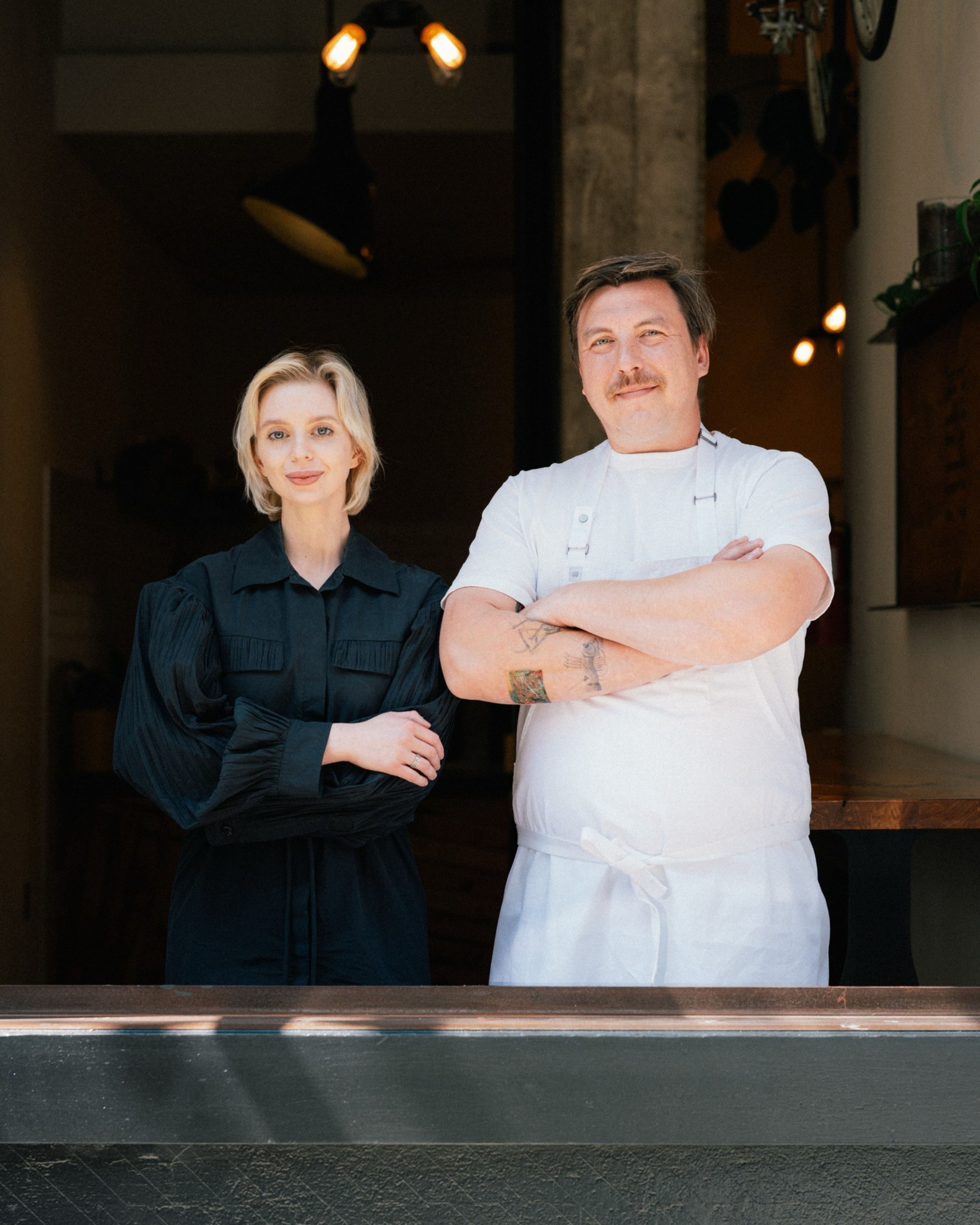 A man in a white shirt and apron stands beside a woman in a black dress. They're in a dimly lit space with light fixtures above them, appearing calm and composed.
