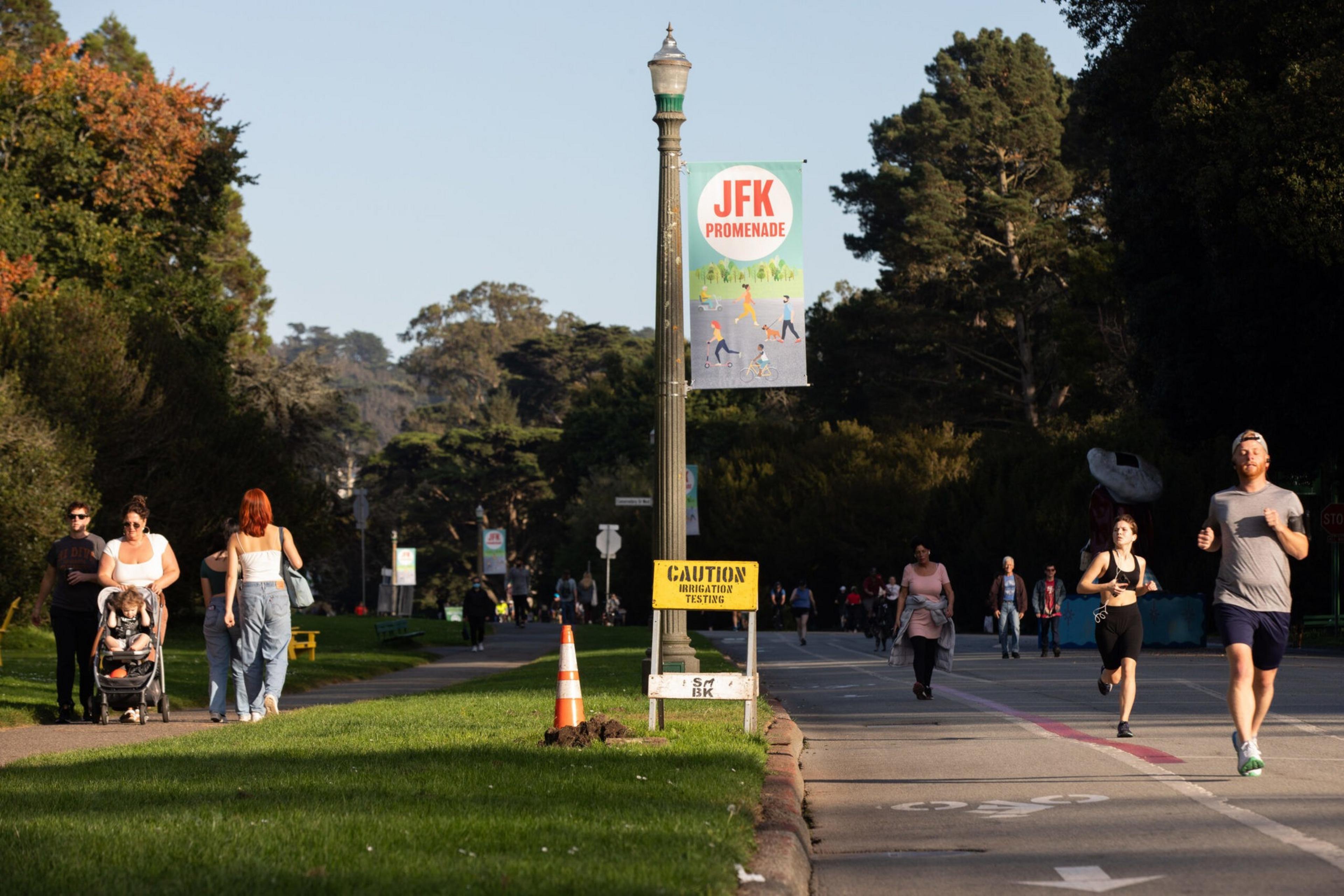 People walk and jog along the JFK Promenade, with trees lining the path and a lamp post displaying signs and a caution notice.