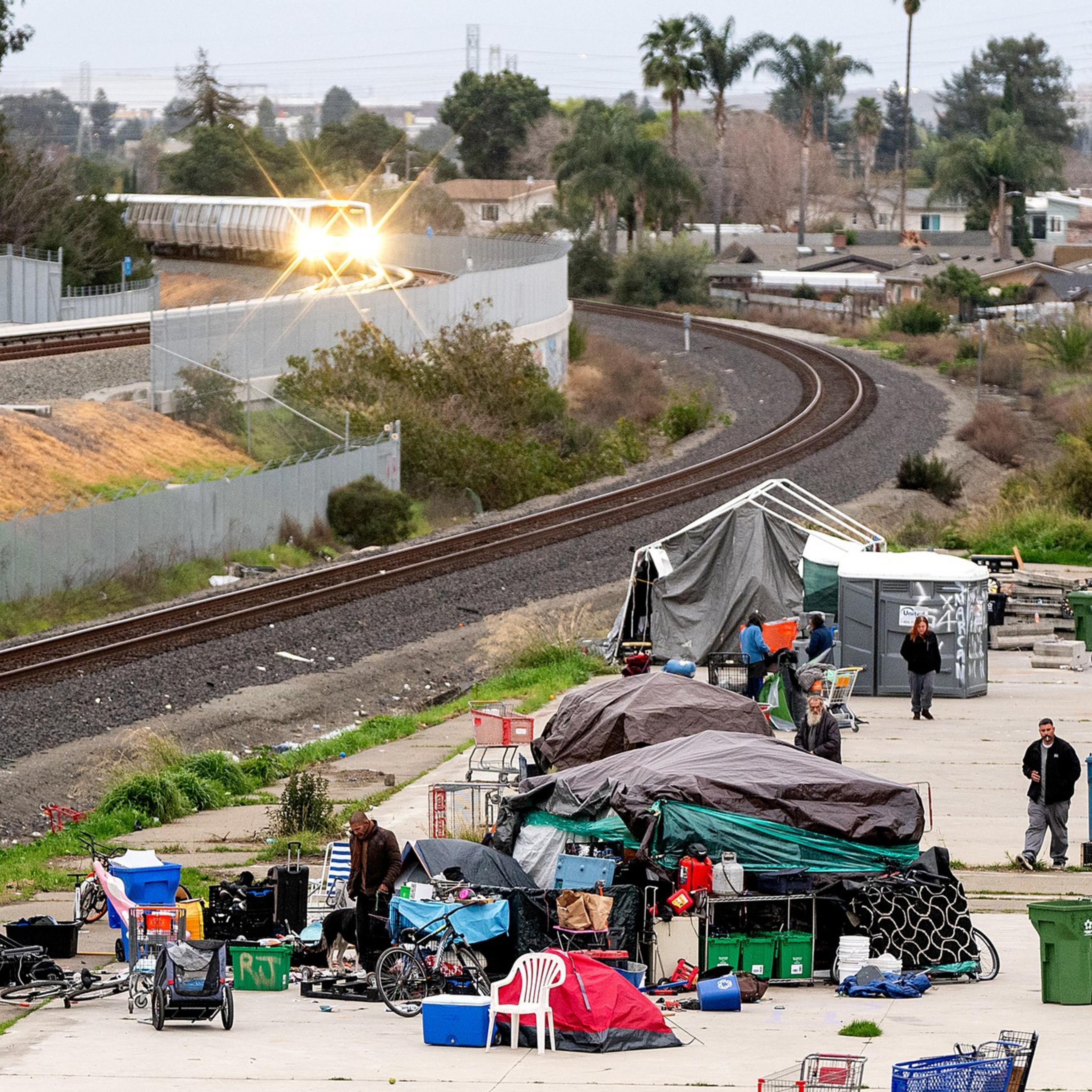 A group of tents and belongings is set up along a curved train track. People are around the makeshift area, with scattered items and several large bins.