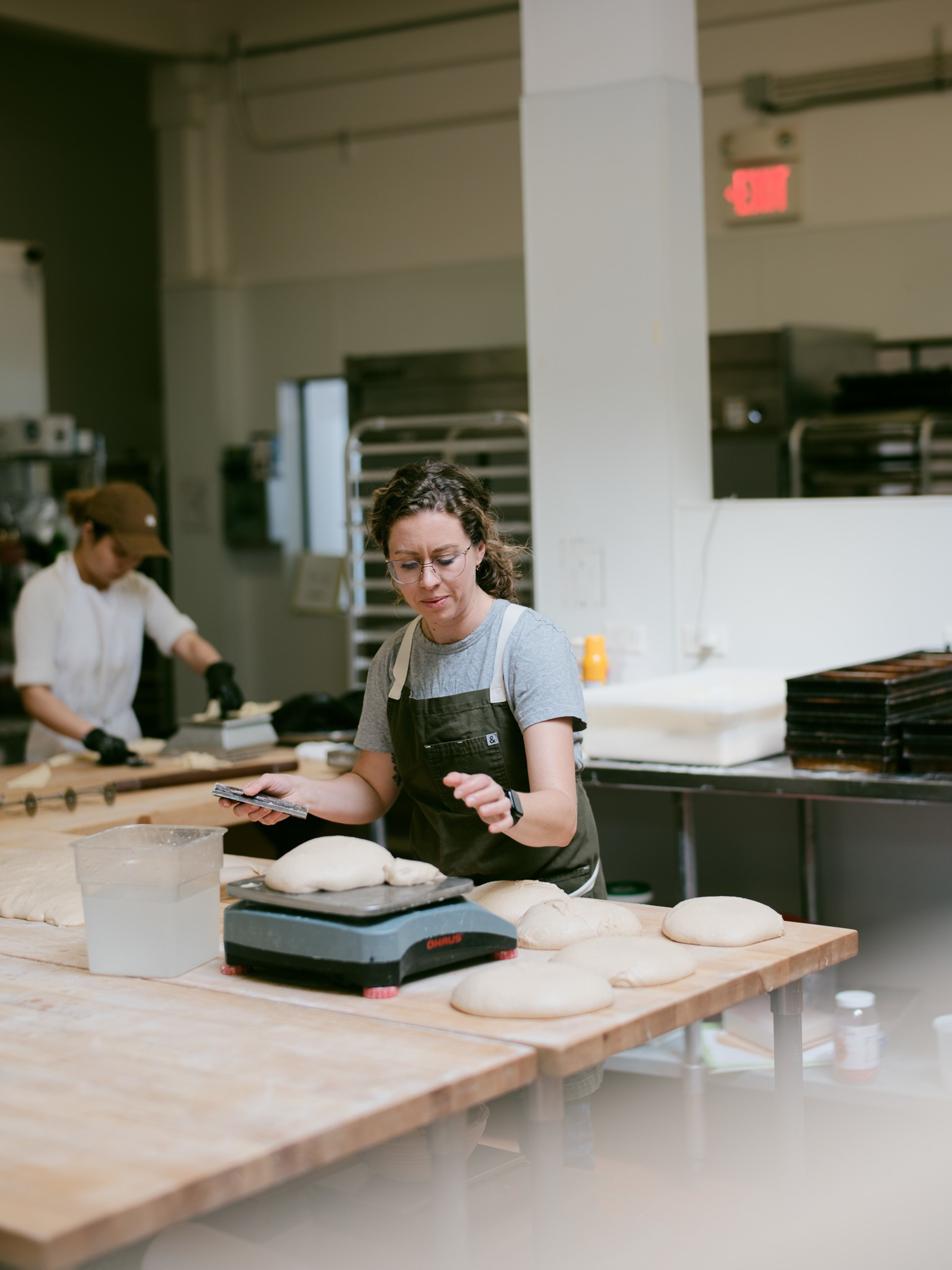 A woman in an apron weighs dough balls on a scale in a bakery kitchen, with more dough and baking trays on the tables around her.
