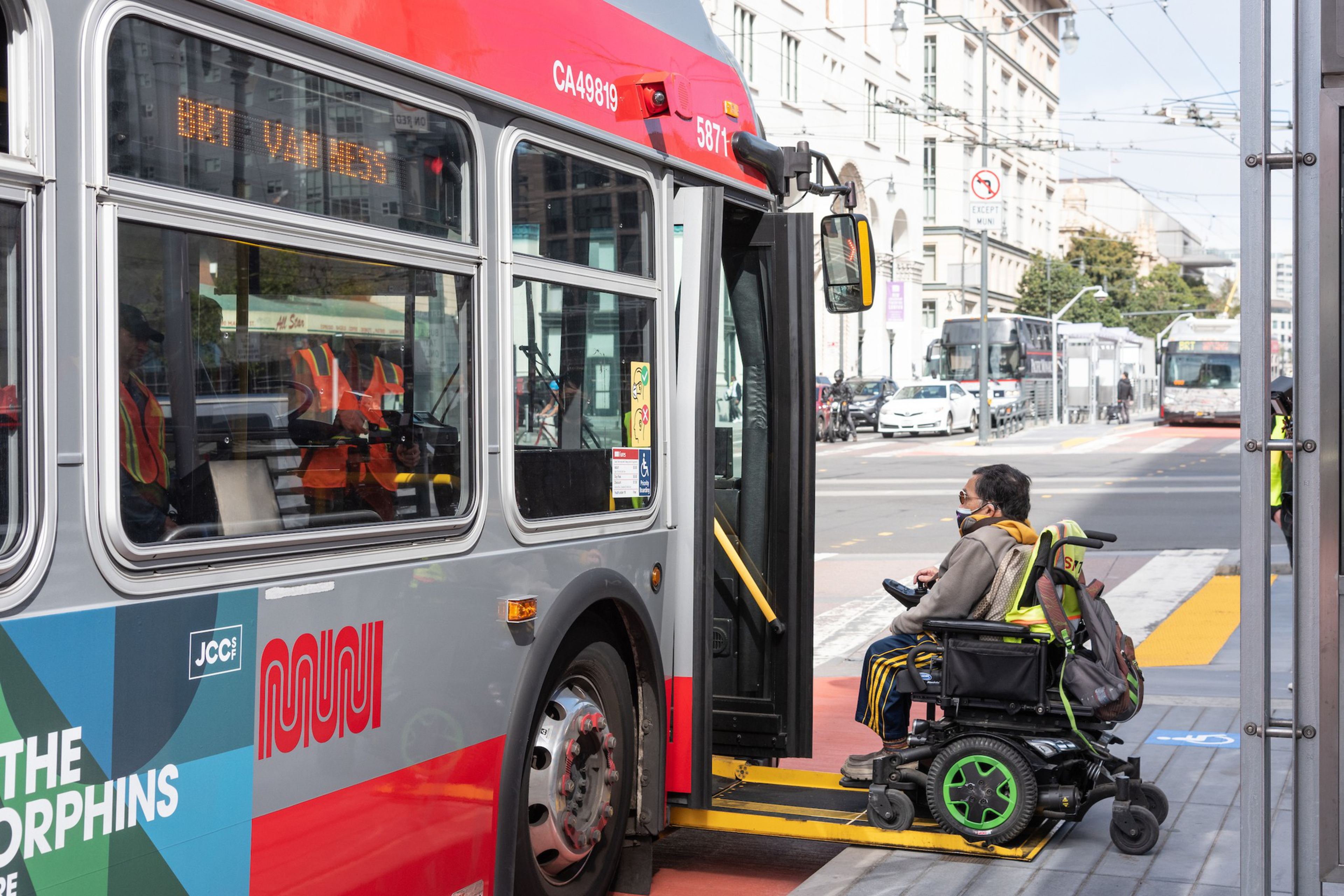 A person in a motorized wheelchair uses a ramp to board a red and gray city bus at a street stop.