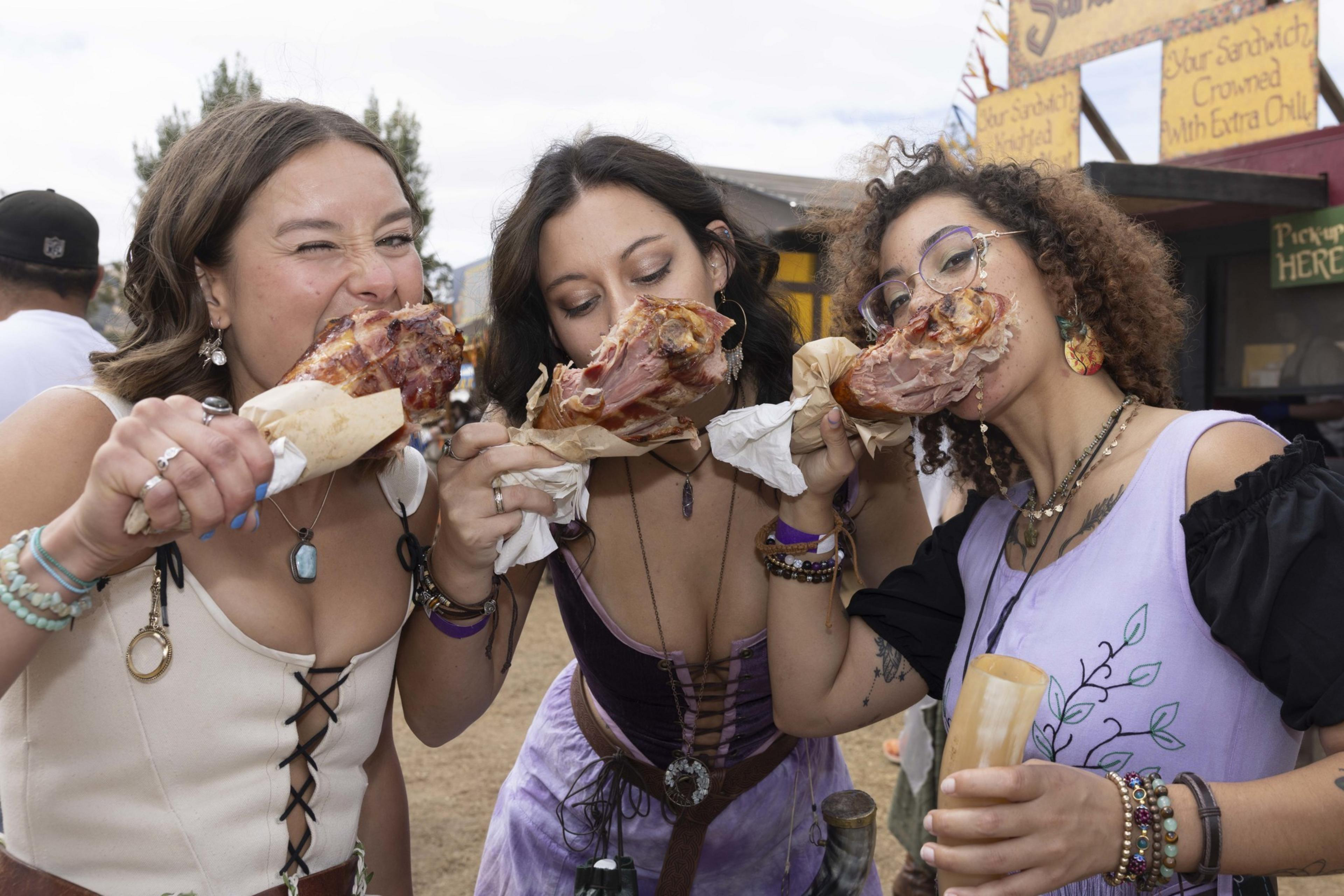 Three women dressed in colorful, Renaissance-style outfits are joyfully biting into large turkey legs at an outdoor festival.