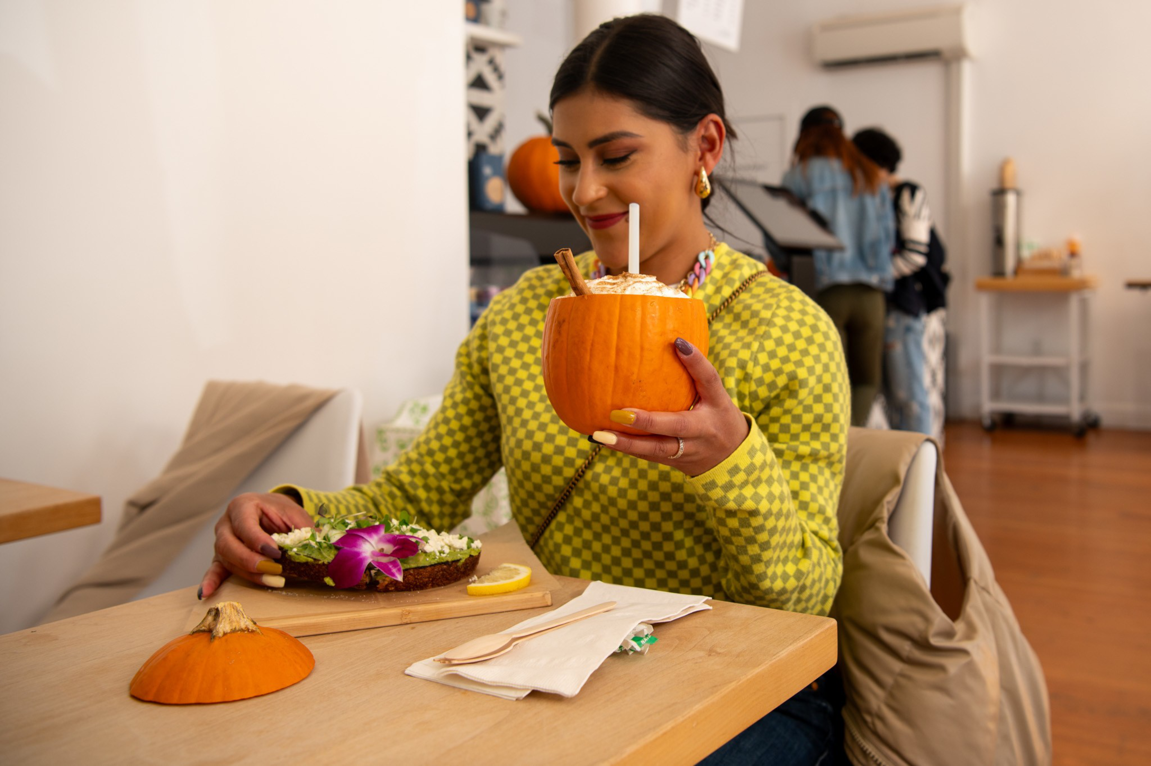 A woman in a yellow checkered sweater holds a whipped cream-topped drink inside a hollowed pumpkin while sitting at a table with a bagel and a pumpkin lid.