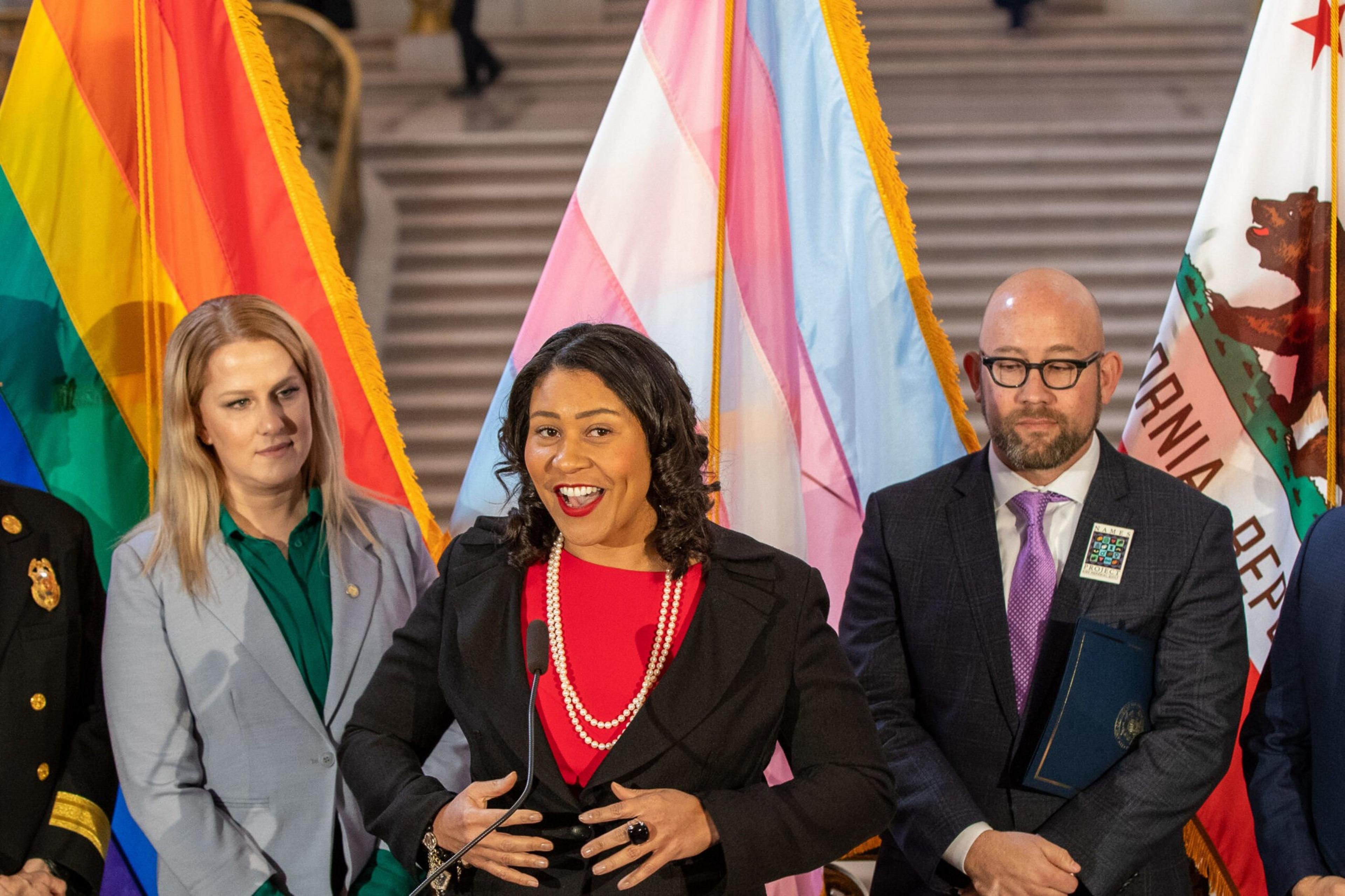 A smiling woman speaking at a podium, flanked by people and colorful pride and trans flags.