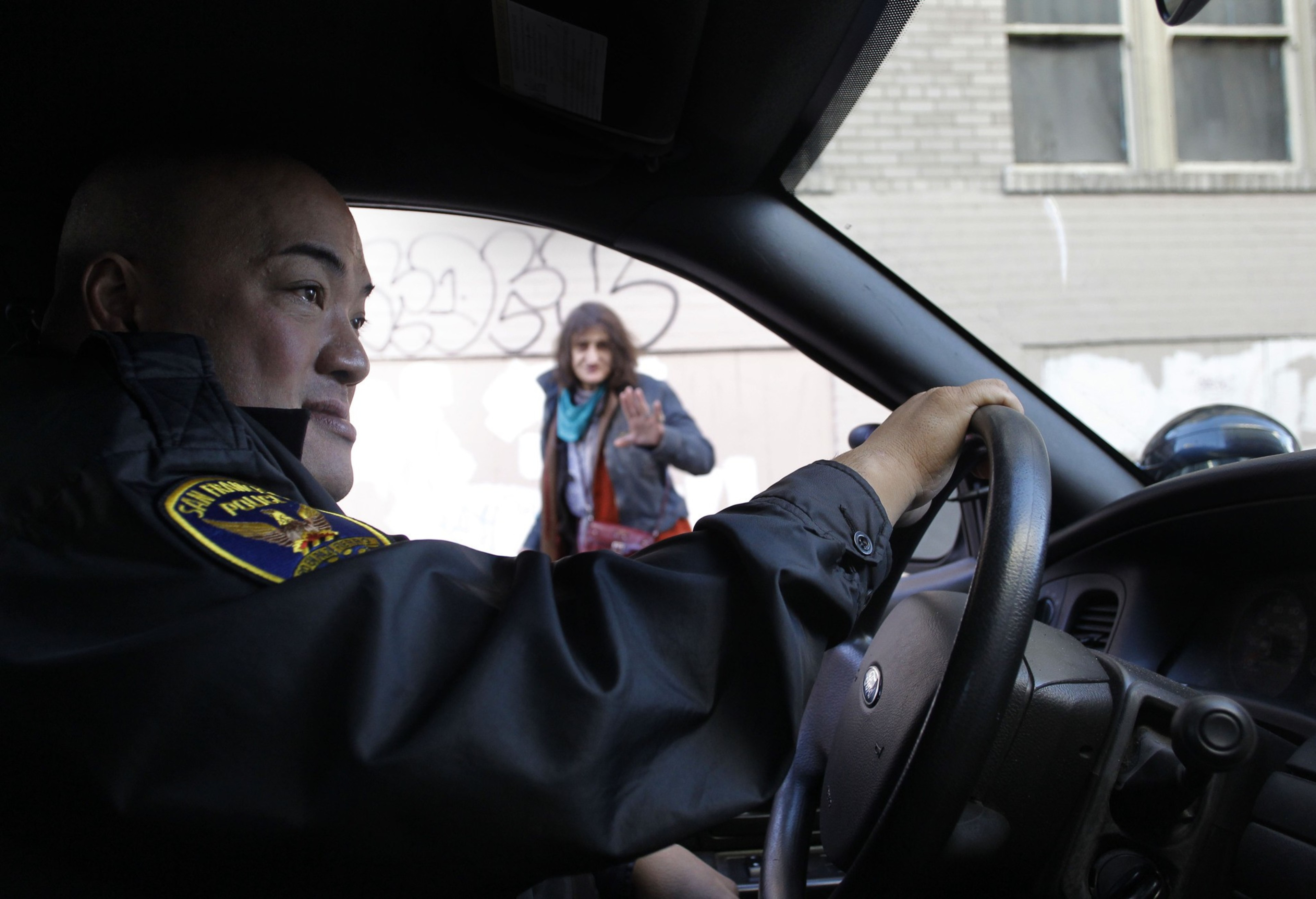 A police officer in uniform sits inside a patrol car, holding the steering wheel, while a woman outside raises her hand and looks toward the car.