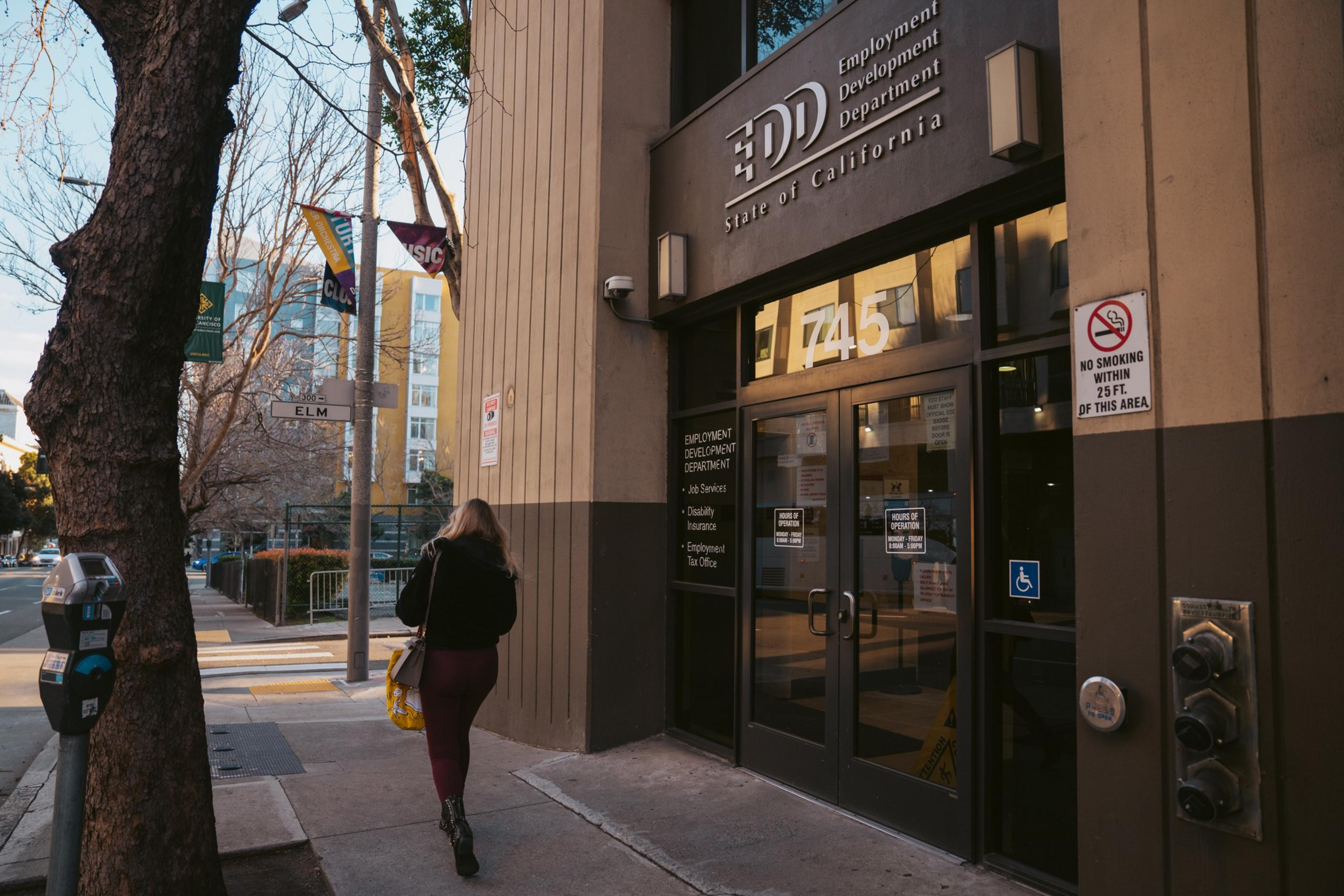 A person walks past the entrance to the California Employment Development Department building at 745 Elm Street, with trees and parking meters nearby.