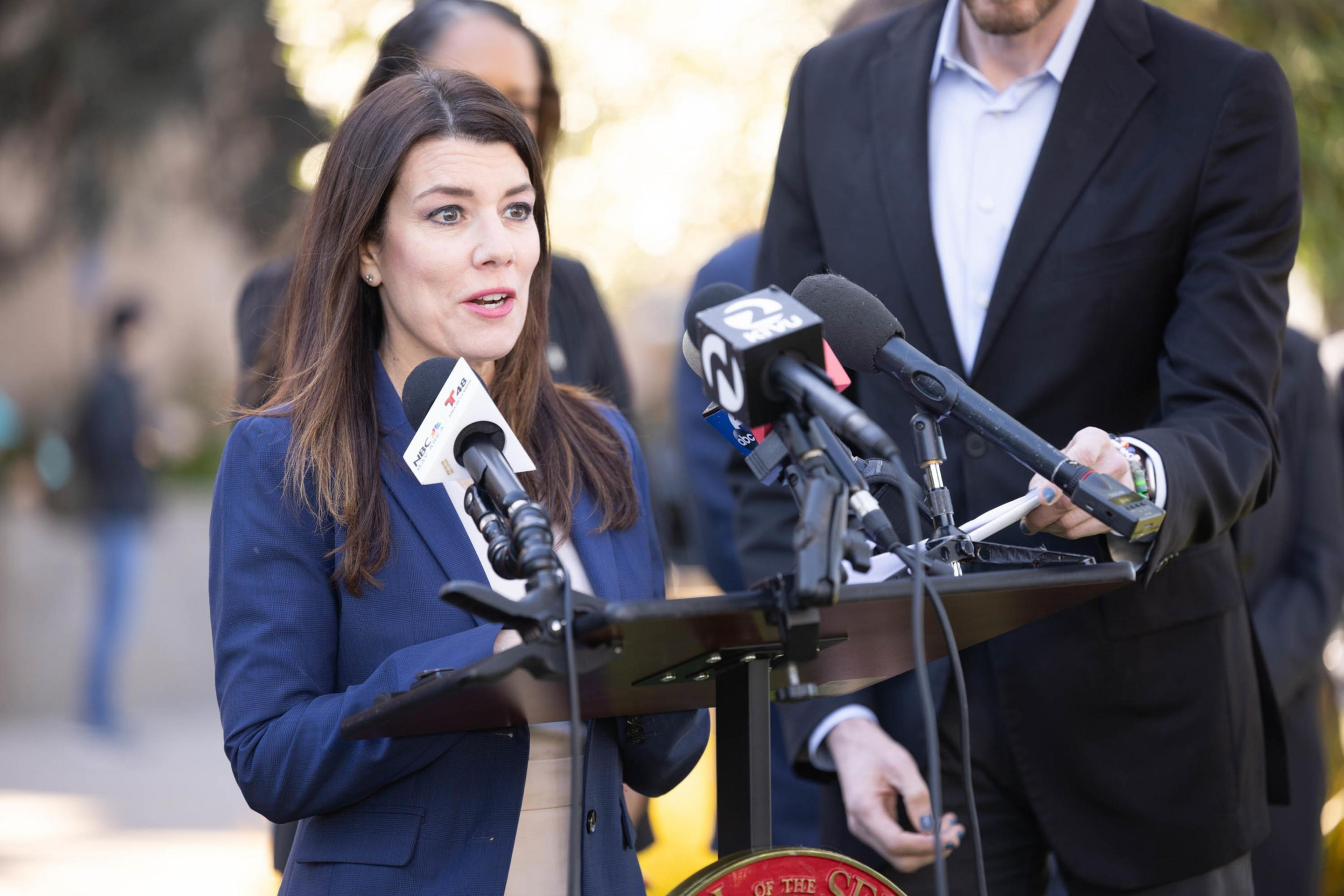 A woman speaks at a podium with microphones, flanked by a man, outdoors with trees in the background.