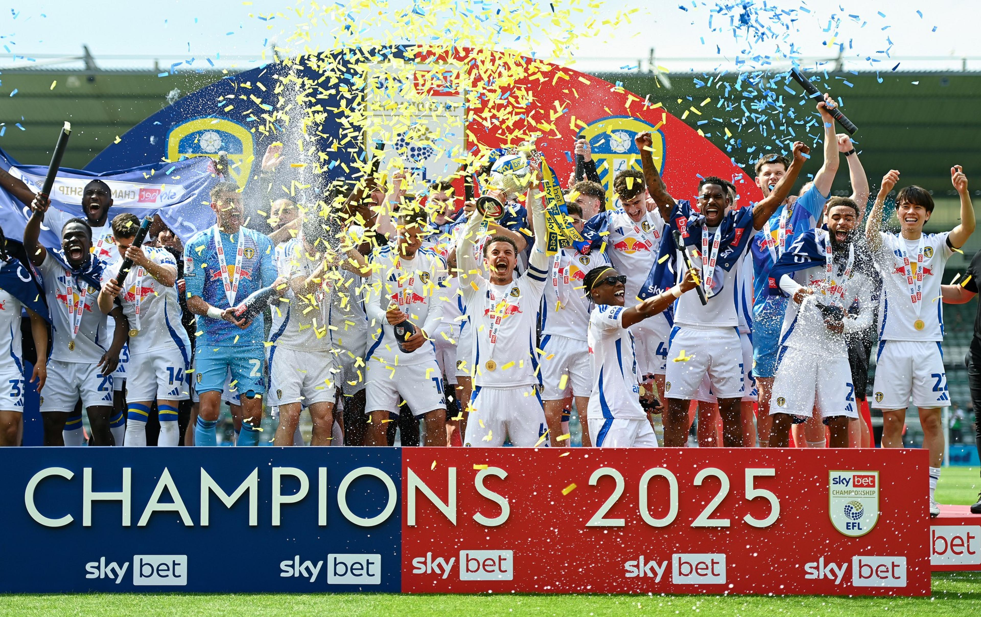 A football team in white uniforms celebrates their 2025 championship win with confetti, medals, trophies, and raised arms on a decorated stage.