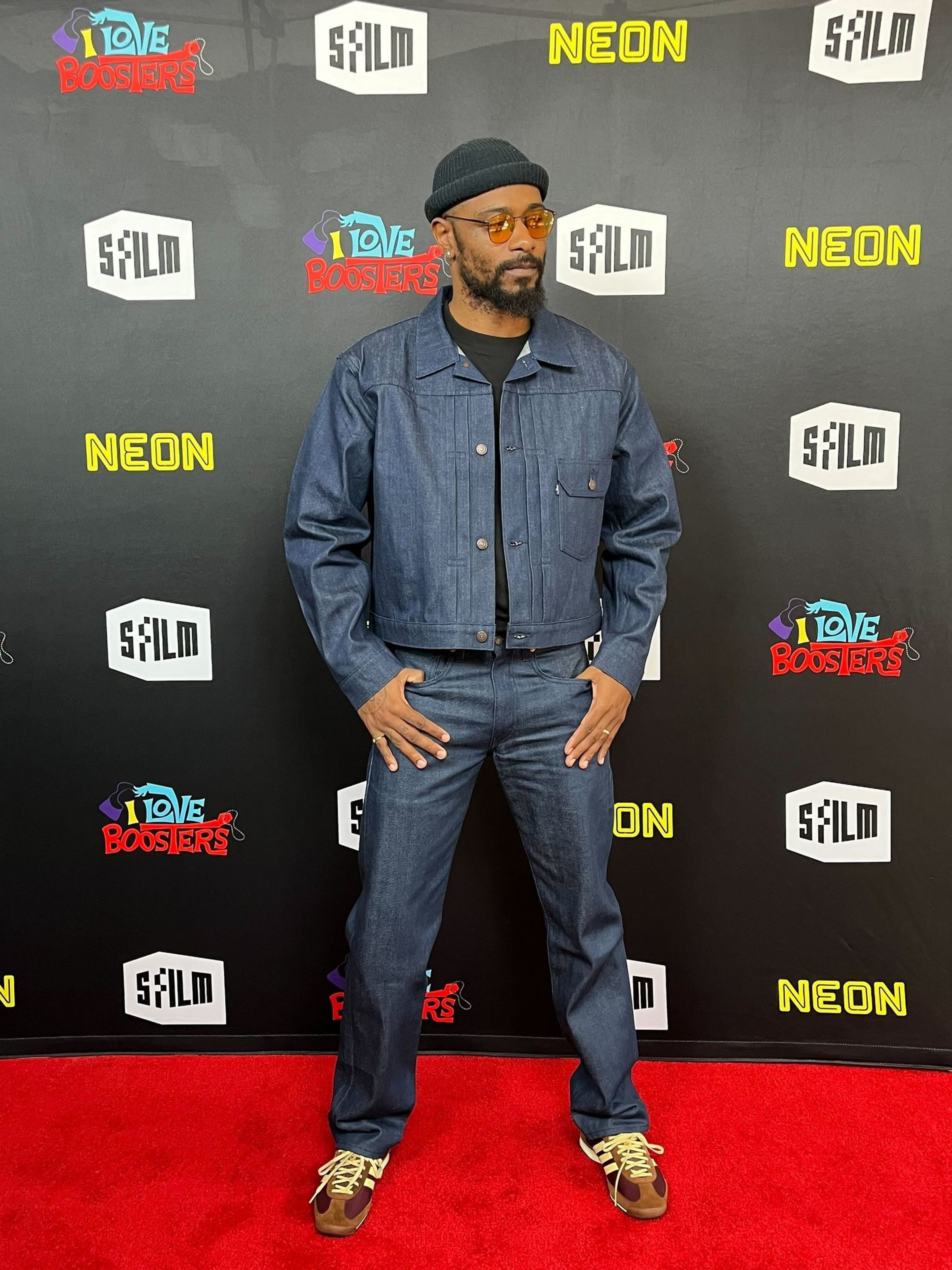 A man stands on a red carpet wearing a dark denim jacket, matching jeans, brown sneakers, round orange sunglasses, and a black beanie.