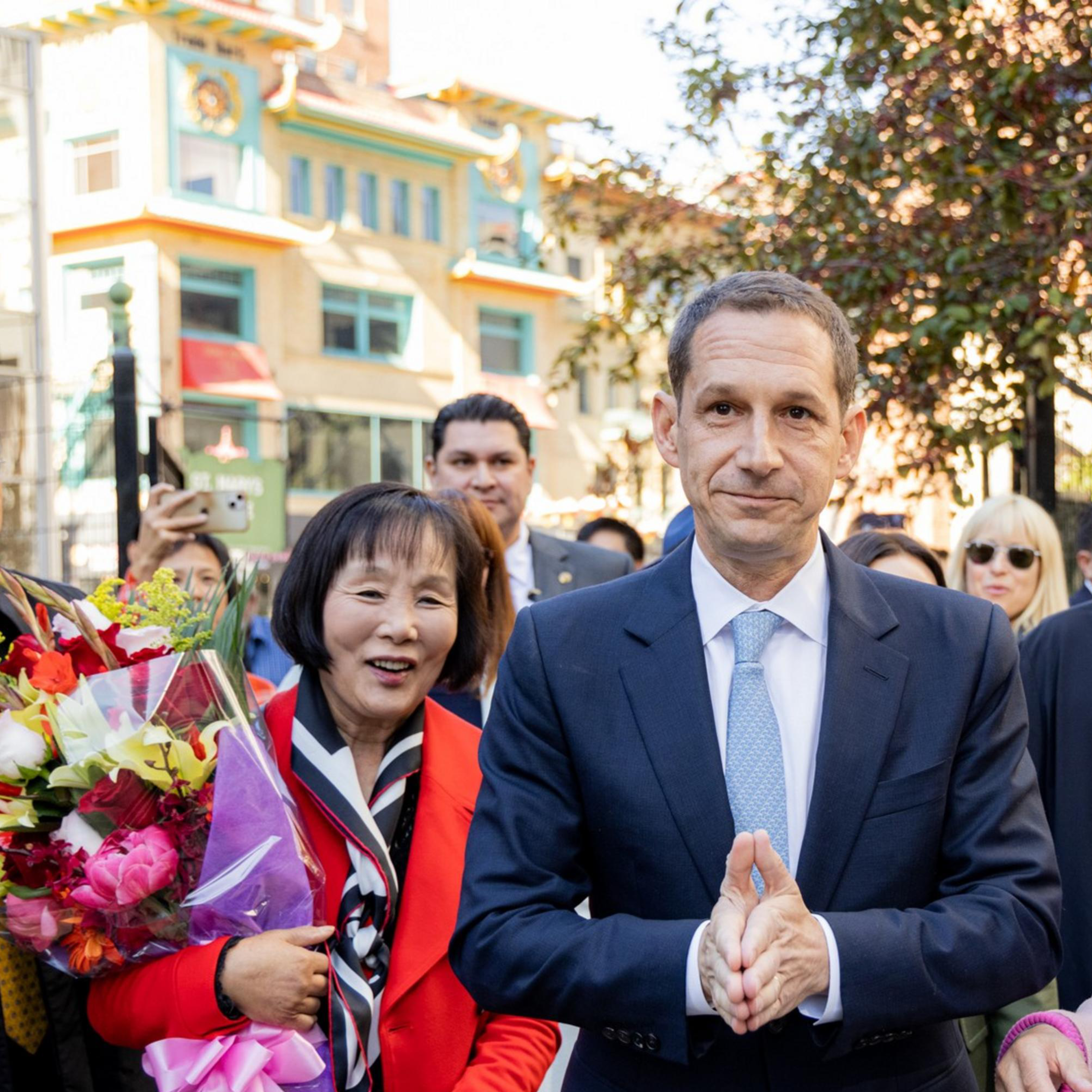 A man in a suit walks surrounded by smiling people; one woman holds a bouquet, and the group seems happy and engaged in conversation.