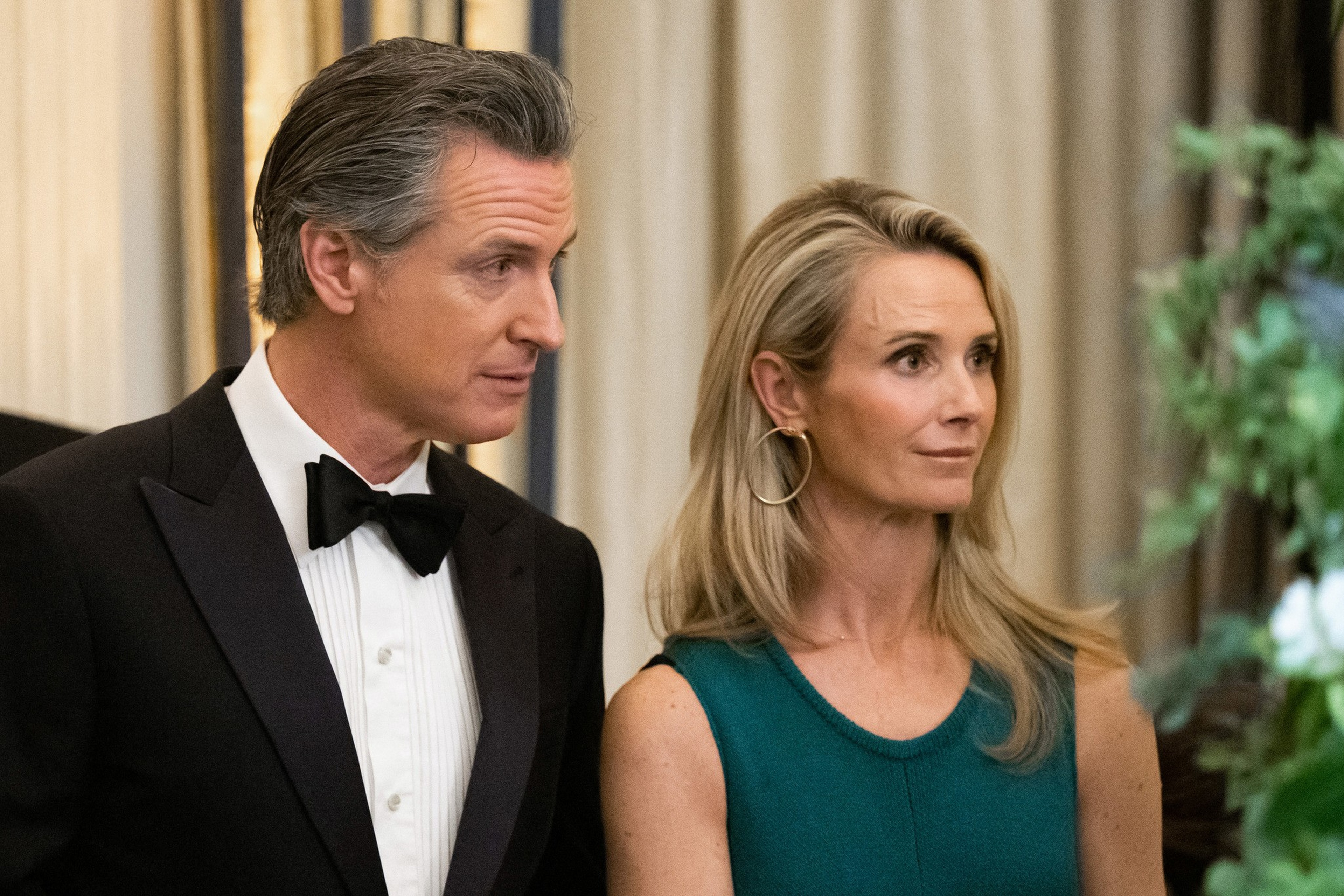 A man in a black tuxedo and bow tie stands next to a woman in a teal sleeveless dress with large hoop earrings, both looking to the right.