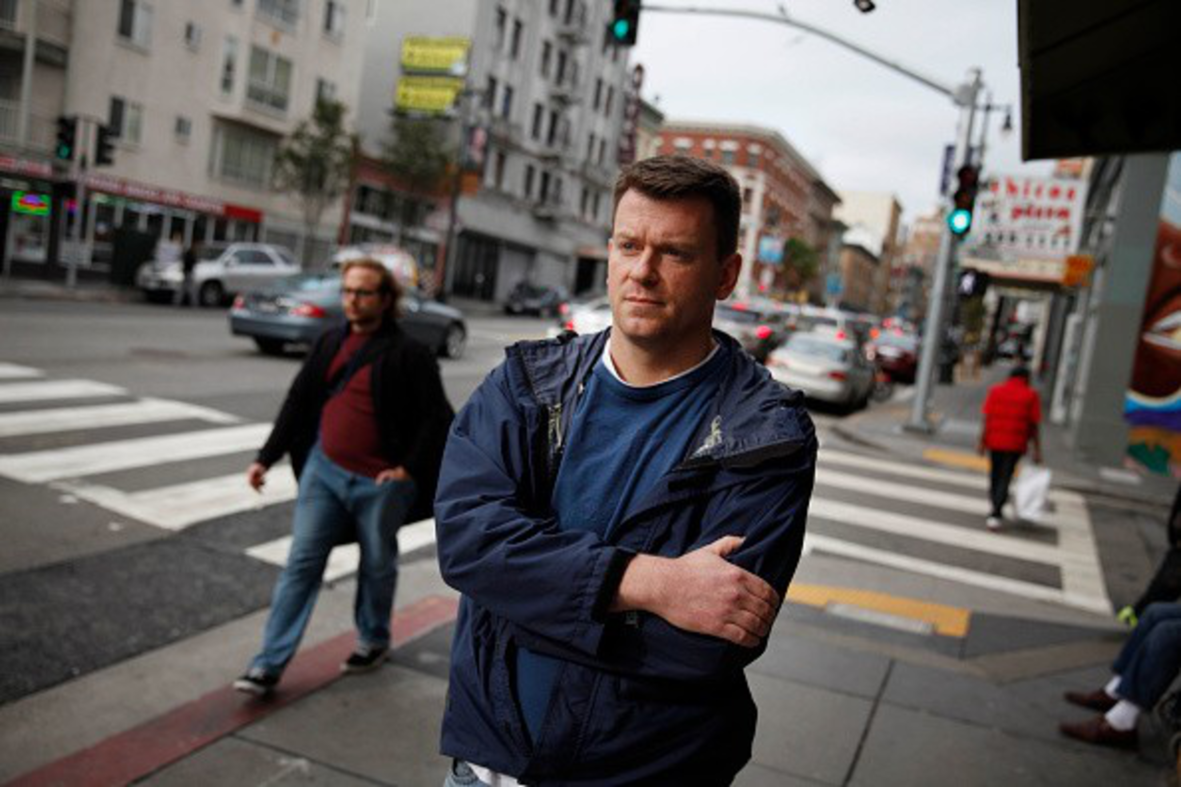 A man in a blue jacket stands on a city sidewalk with arms crossed, looking serious, while people and cars move in the busy street background.