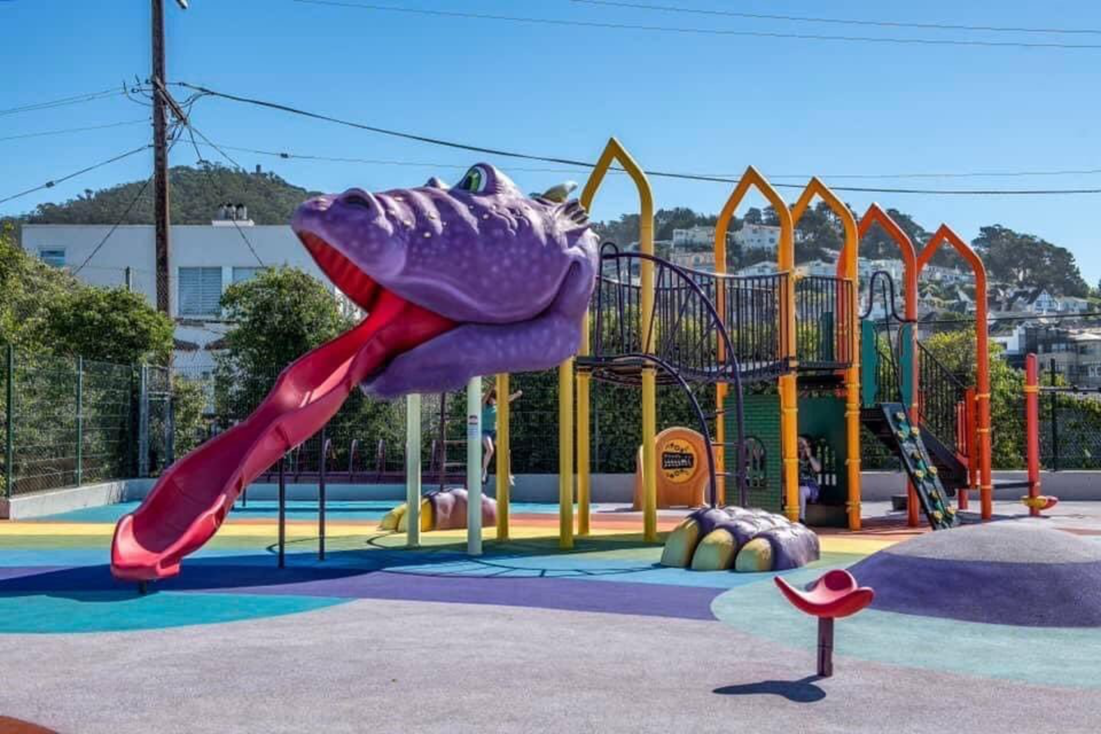 A colorful playground features a large purple dragon sculpture with its tongue forming a slide, surrounded by climbing structures and multi-colored poles.